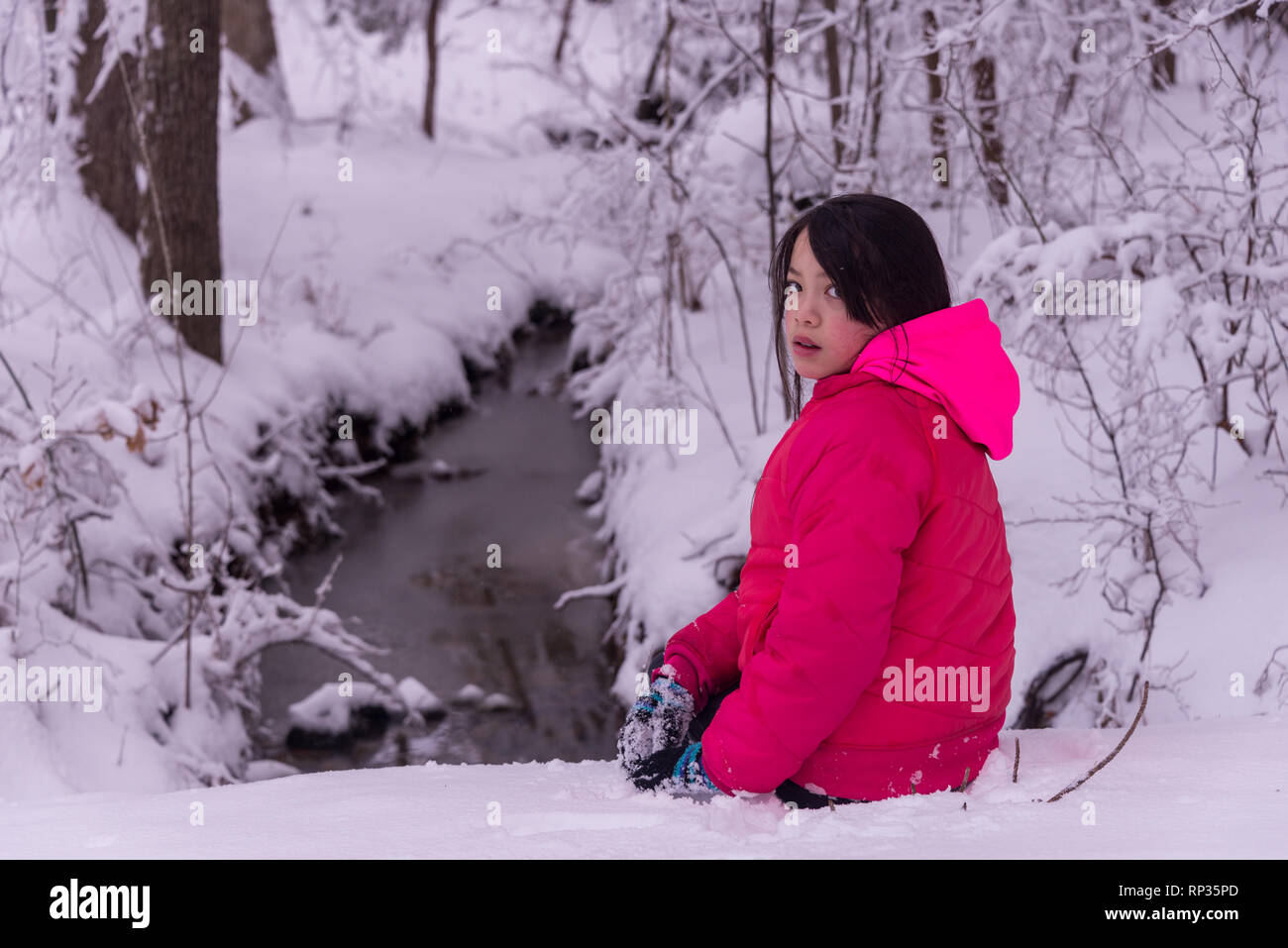 Junge asiatische Mädchen sitzen im Schnee vor Stream Stockfoto