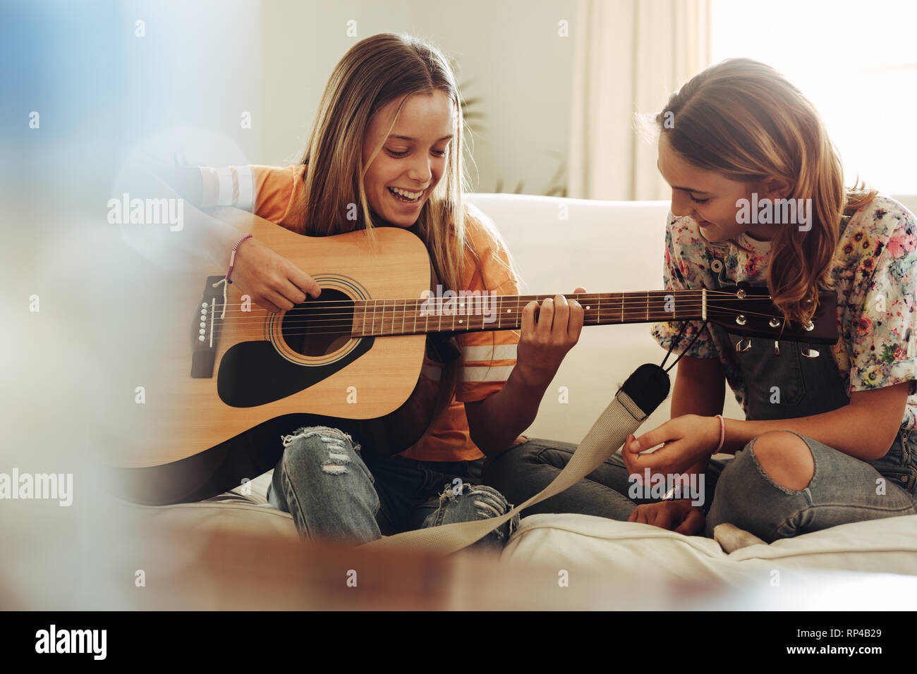 Fröhlicher junger Mädchen, Gitarre zu spielen, zu Hause zu sitzen. Mädchen spielen Musik auf Gitarre, während ihre Freunde auf aussieht. Stockfoto