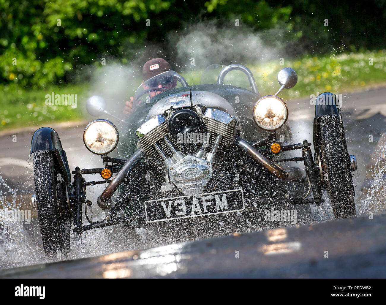 Morgan 3-wheeler Spritzwasser durch eine Furt an Geschwindigkeit mit Scheinwerfern und Reflexionen in der nassen Straße. Model Release Form unterzeichnet. Stockfoto