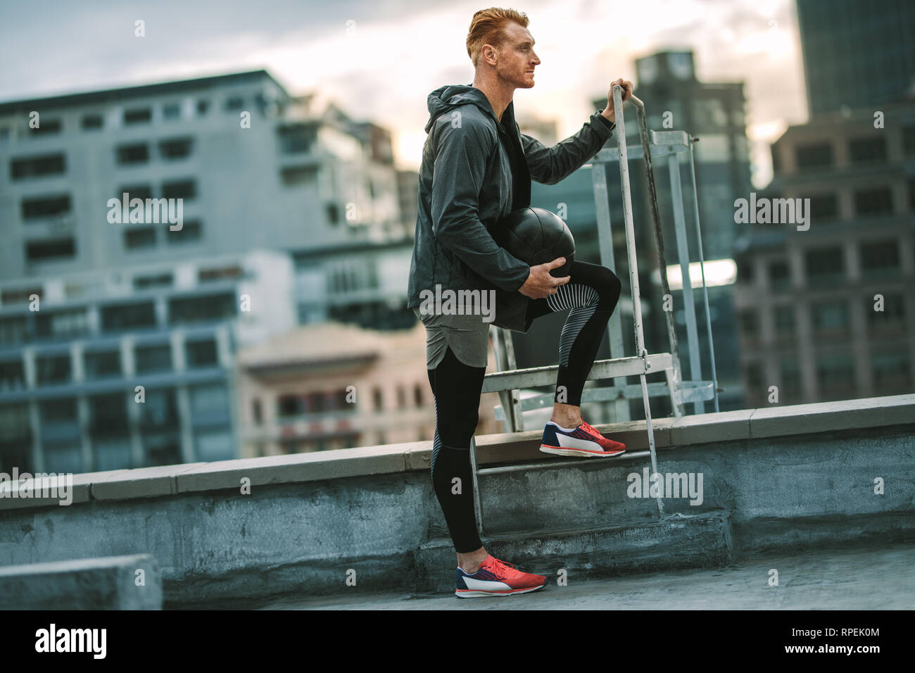 Athlet steht in der Nähe eine Dachterrasse mit einem Treppenaufgang mit einem medizinball in der Hand. Seitenansicht eines Mannes in fitness Verschleiß auf der Dachterrasse zu tun Fitness Training. Stockfoto