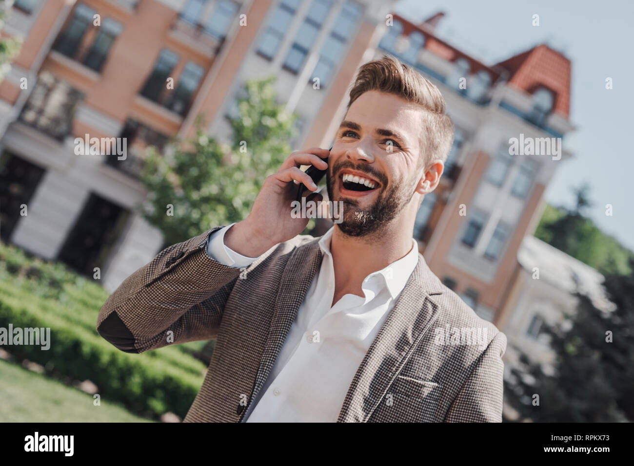 Portrait Of Happy Geschäftsmann Walking im Freien mit Handy. Mann mit Handy. Glückliche Menschen sprechen. Unternehmer im Freien mit Telefon. Er trägt smart casual Outfit der braunen Sakko und weißem T-Shirt. Stockfoto