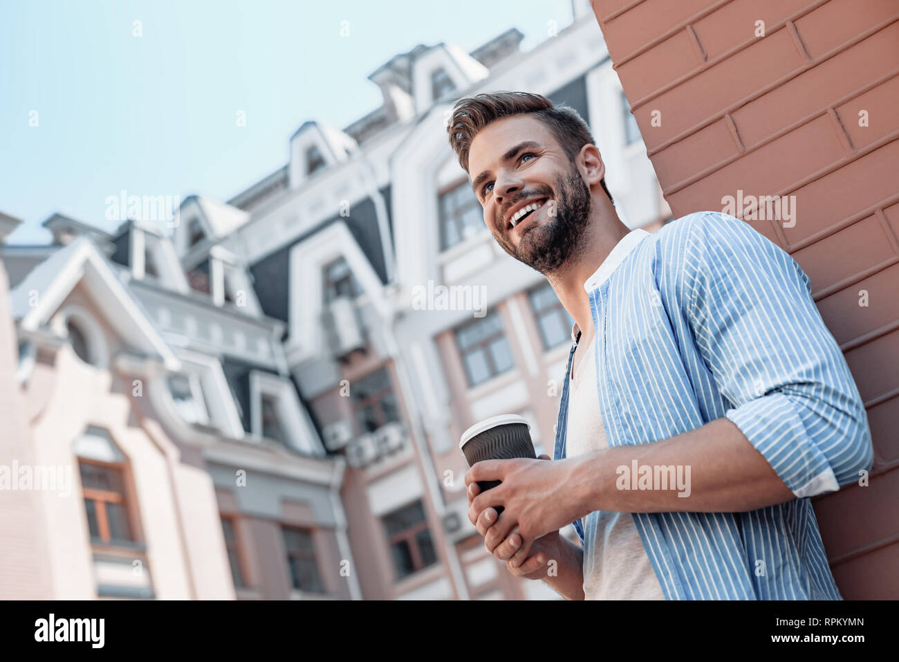 Junge stilvollen Mann mit blauem Hemd und grauen T-Shirt stehen auf der Straße mit einer Tasse Kaffee neben der Suche und lächelt fröhlich. Unscharfer Hintergrund Stockfoto