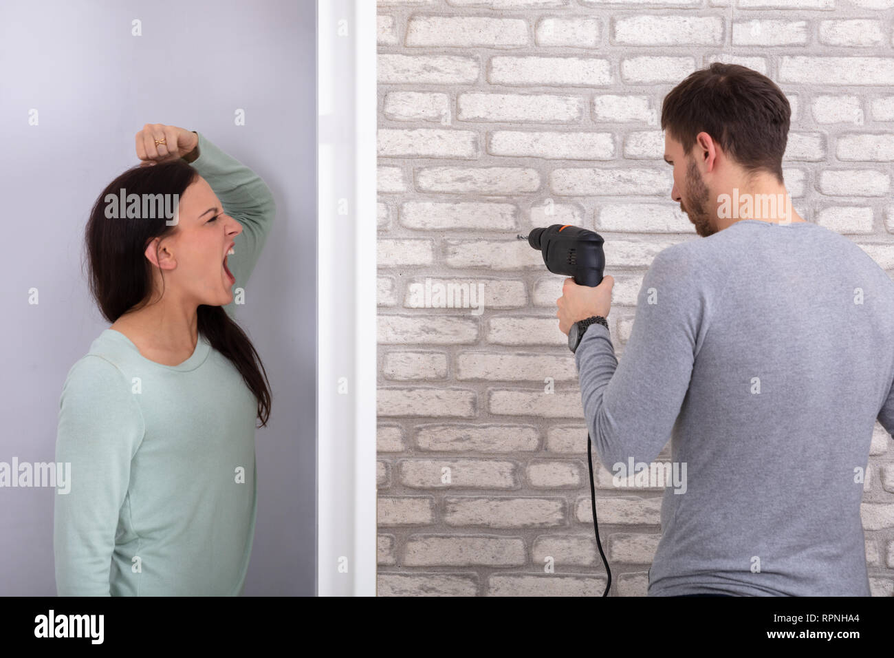 Frau schreiend hinter der Wand mit Nachbarn Mann mit Bohrmaschine auf weißer Wand Stockfoto
