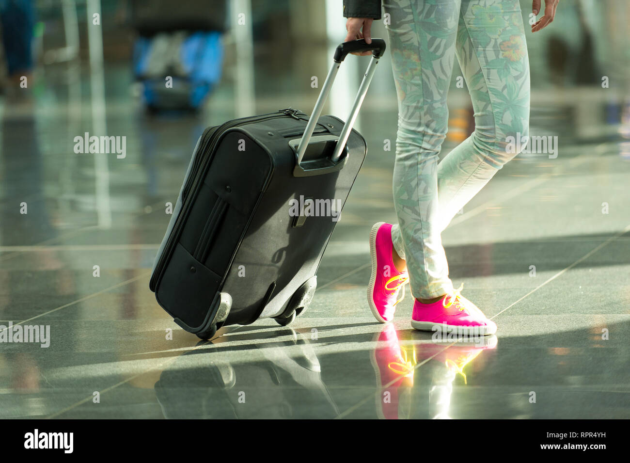 Schwarze Reisetasche oder Koffer auf Rädern in der Hand und weibliche Beine in Jeans und leuchtend rosa Gumshoes auf Füße auf grauem Fliesenboden. Reisen und Gepäck. Ferienhäuser und Mode Stockfoto