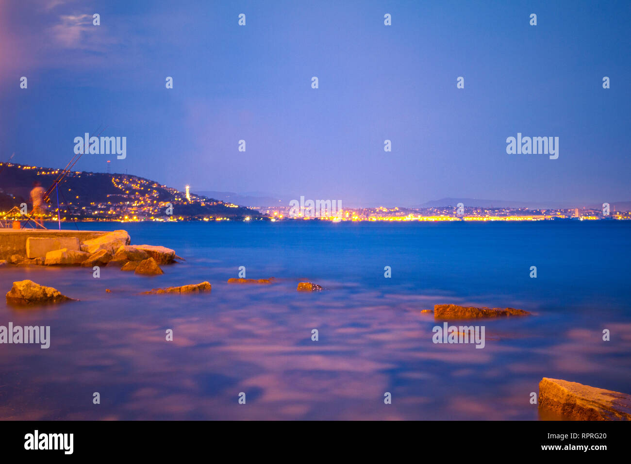 Vittoria Licht, Triest in der Nacht. Blau und Violett, Dämmerung, City Lights, Adria Küste, Felsen, weit entfernten Leuchtturm. Lebendige Farben. Lange Belichtung Stockfoto