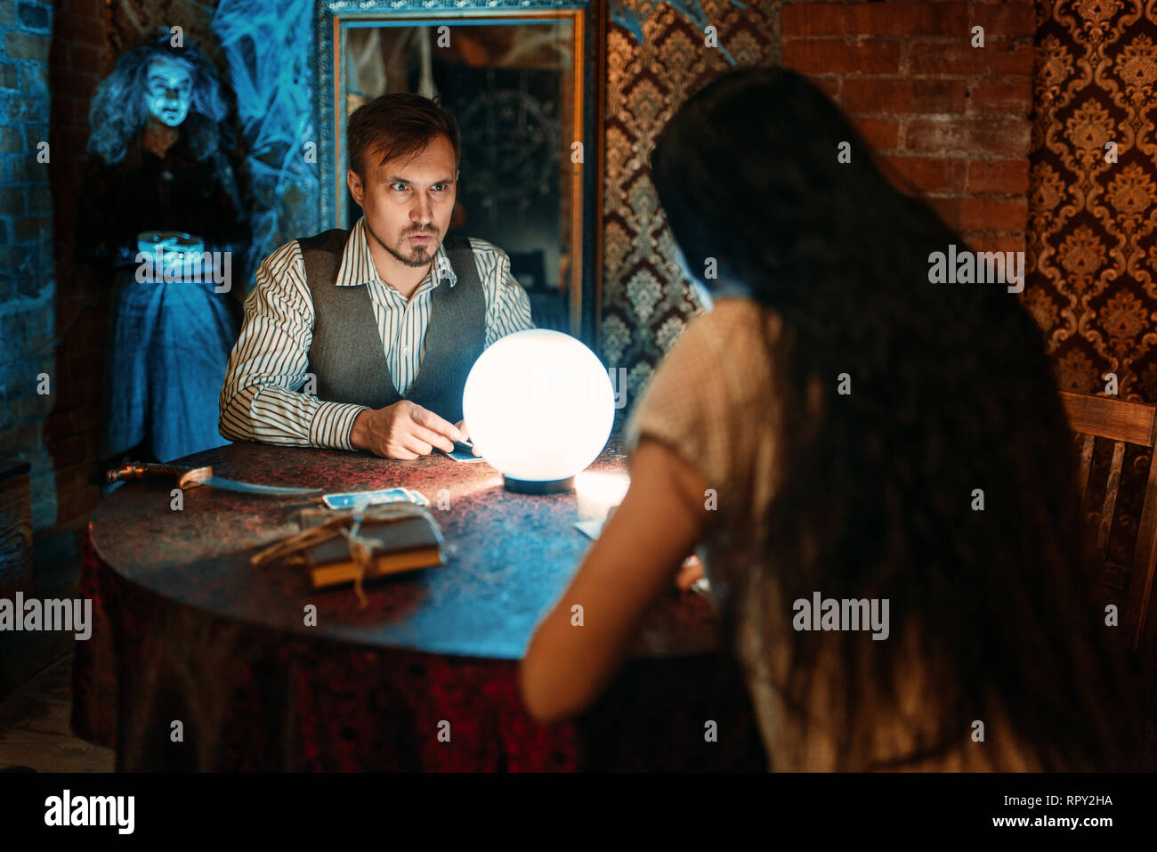 Junge Leute am Tisch mit Crystal Ball auf spirituelle Seance, gruselige Hexe auf Hintergrund. Weibliche foreteller ruft die Geister Stockfoto