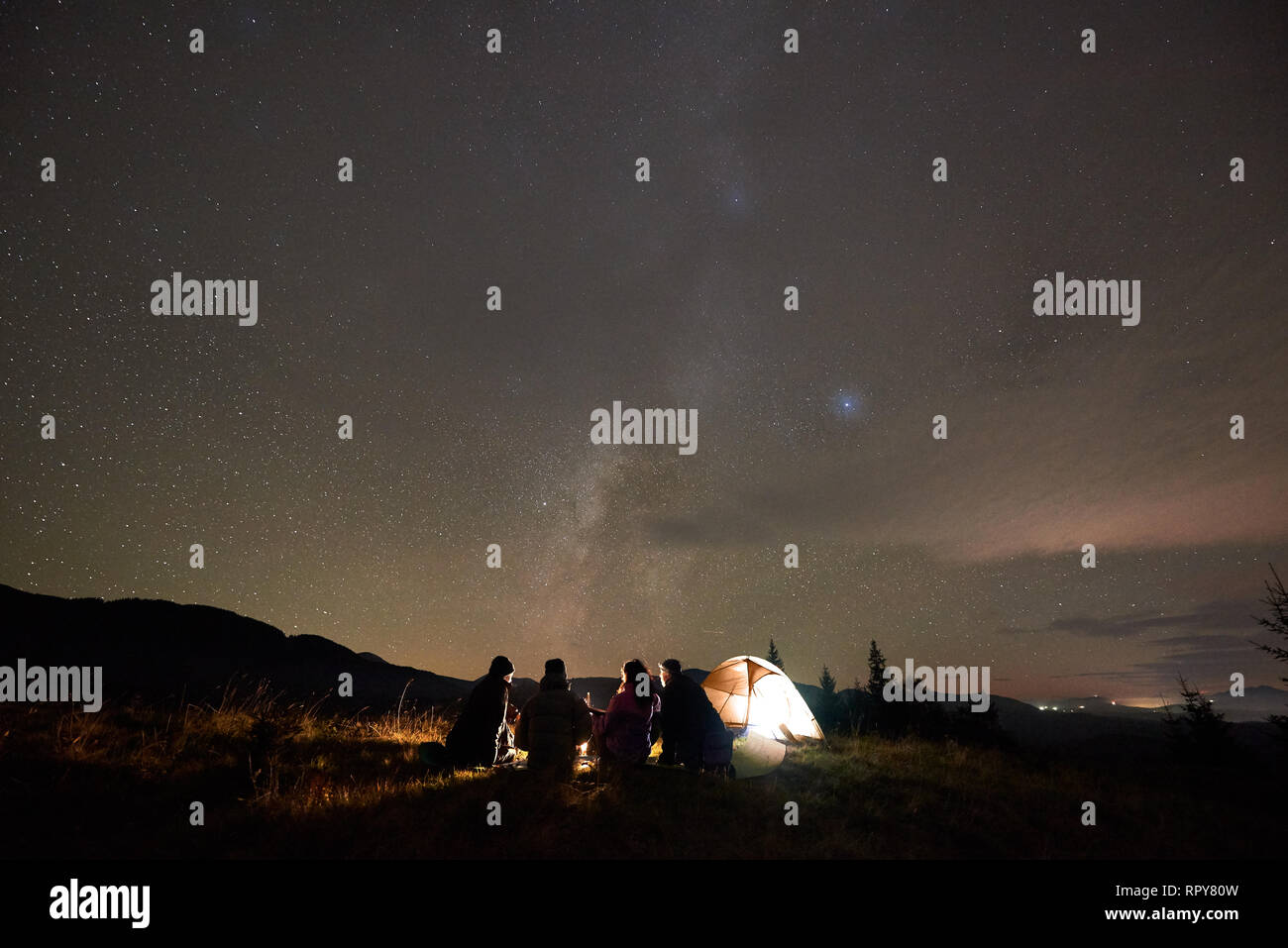 Nacht Camping unter den Sternen, Tourismus und aktiven Lebensstil. Rückansicht Silhouette von vier Personen sitzen auf grasbewachsenen Senke an touristische Hütte und Brennen cam Stockfoto
