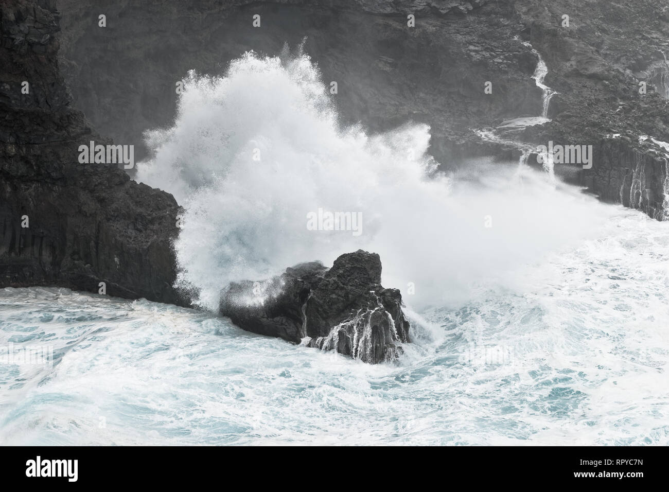 Eine Welle bricht bei stürmischem Wetter auf einer felsigen Küste mit einem kleinen Bucht, Gischt spritzt weit oben, aus dem Felsen, Wasser läuft wieder zurück ins Meer - Ort: Spa Stockfoto