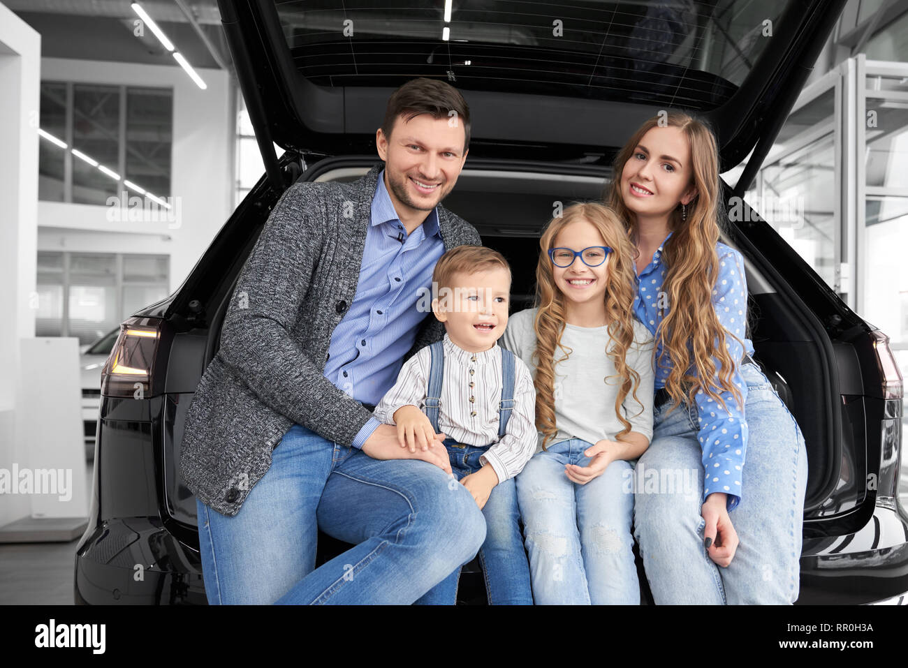 Fröhliche und glückliche Familie zusammen im Auto center Showroom posieren. Eltern und Kinder im Kofferraum sitzen, Platz für Gepäck von schwarzen Automobil auf Kamera und lächelnd. Stockfoto