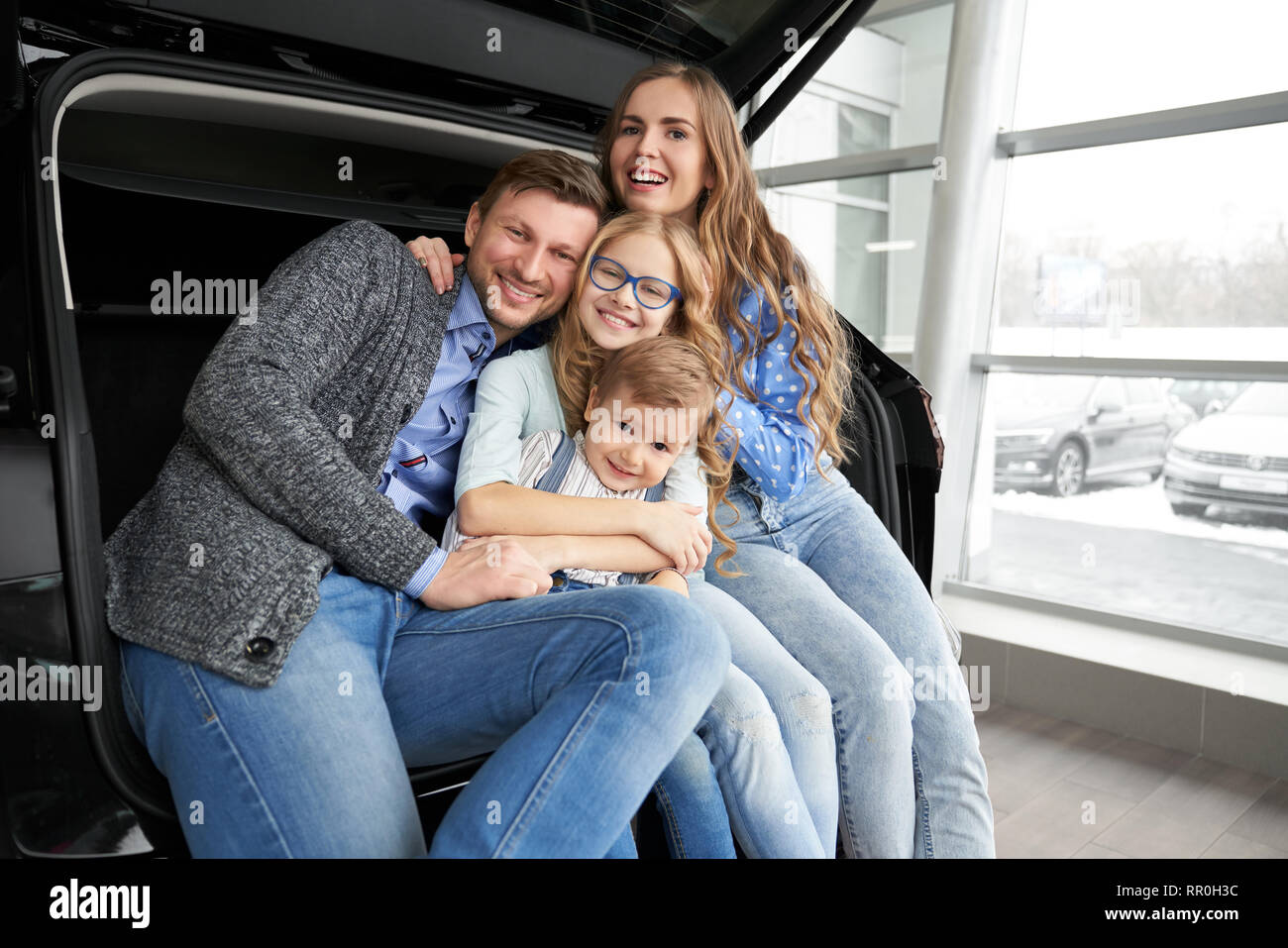 Ziemlich Familie sitzt im Kofferraum von schwarzen Automobil und posieren. Personen, die das Fahrzeug. Glückliche Eltern umarmt Kinder, Sohn und Tochter, von der Kamera schauen und lächeln. Stockfoto