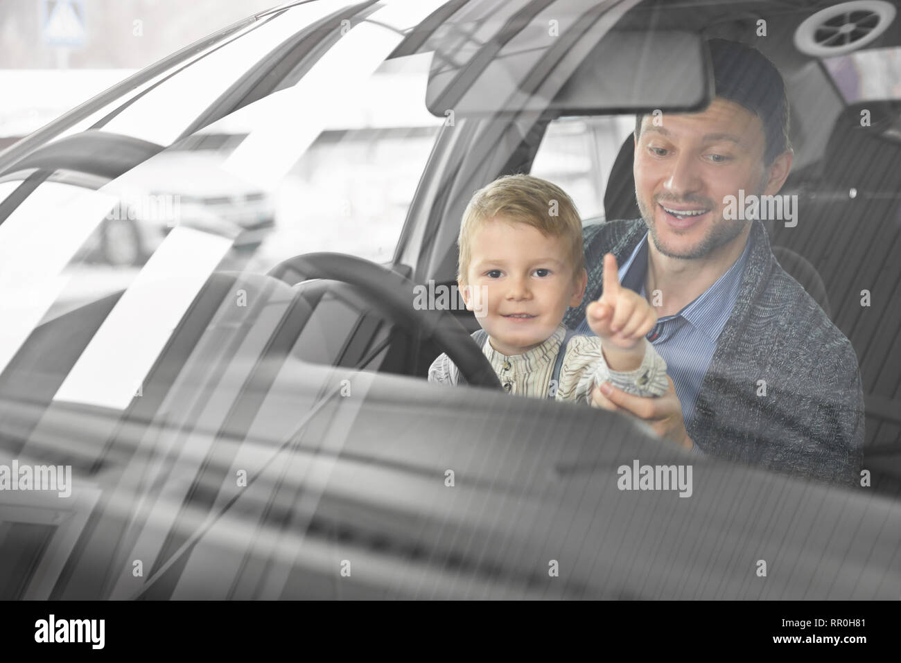 Ansicht von vorne durch die Windschutzscheibe von Vater und Sohn sitzen im Fahrersitz von Automobil- und beobachten Auto Kabine. Kleinen Niedlichen jungen Blick auf Kamera und zeigt mit dem Finger. Stockfoto