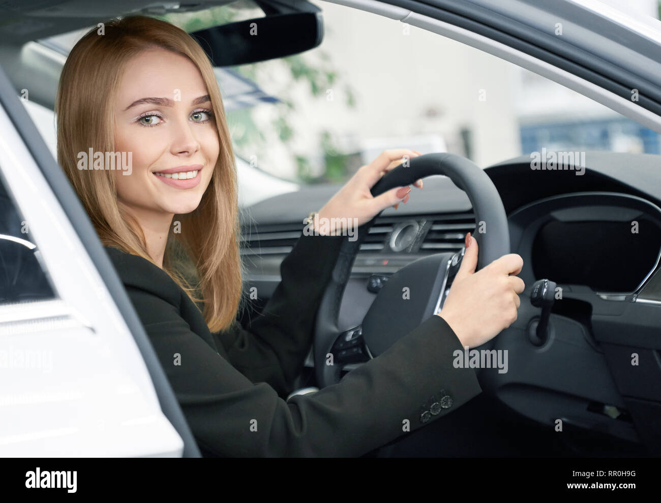 Charmante Frau mit blonden Haaren sitzen in der teuren, neuen Automobil, in die Kamera schauen und lächeln. Potenzielle Käufer des Fahrzeugs testen und beobachten Auto Kabine im Auto im Showroom. Stockfoto