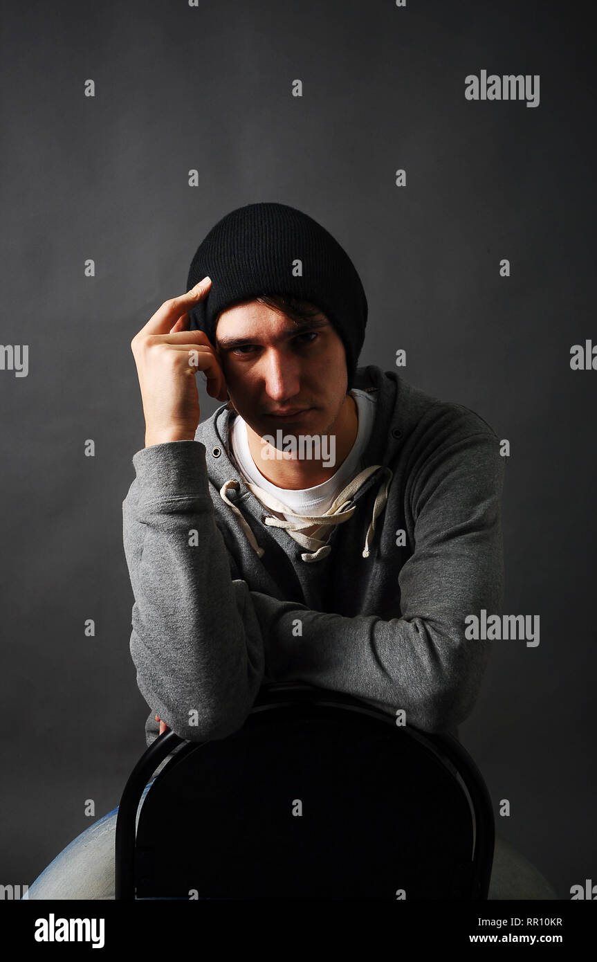 Der junge Mann auf einem Stuhl sitzend in einem grauen T-Shirt und einen schwarzen Hut, seine Hand auf den Kopf im Schatten. Stockfoto