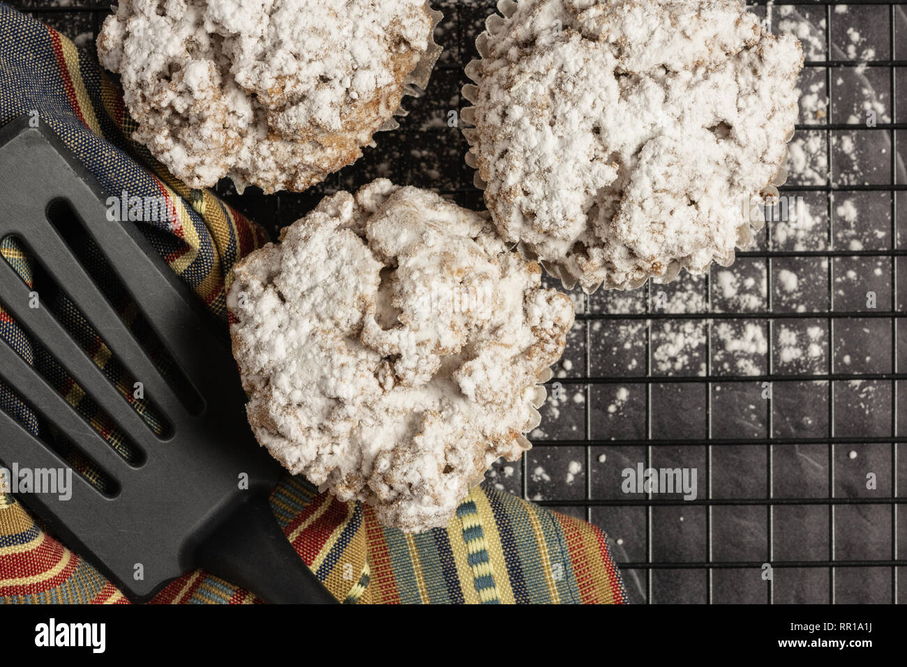 Drei Zimt crumb Muffins und einen gedämpften Farben Streifen Serviette mit Teigschaber in einem Rack mit Puderzucker auf dem schwarzen Marmor Arbeitsplatte Stockfoto