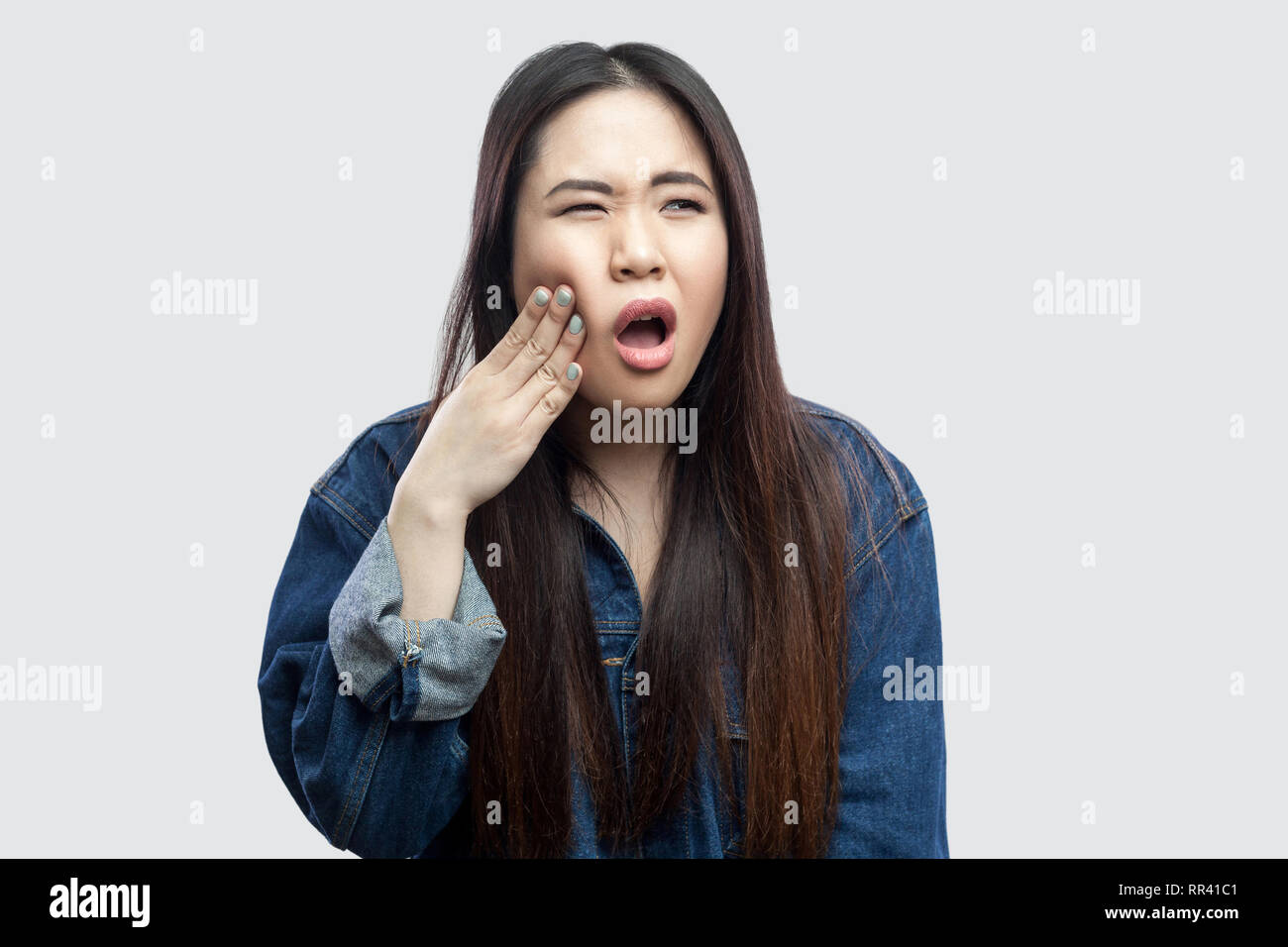 Zahn weh. Portrait von Sick schöne Brünette asiatische junge Frau in lässigem Blue Denim Jacke mit Make-up stehen und ihren schmerzlichen Zahn Holding. indo Stockfoto