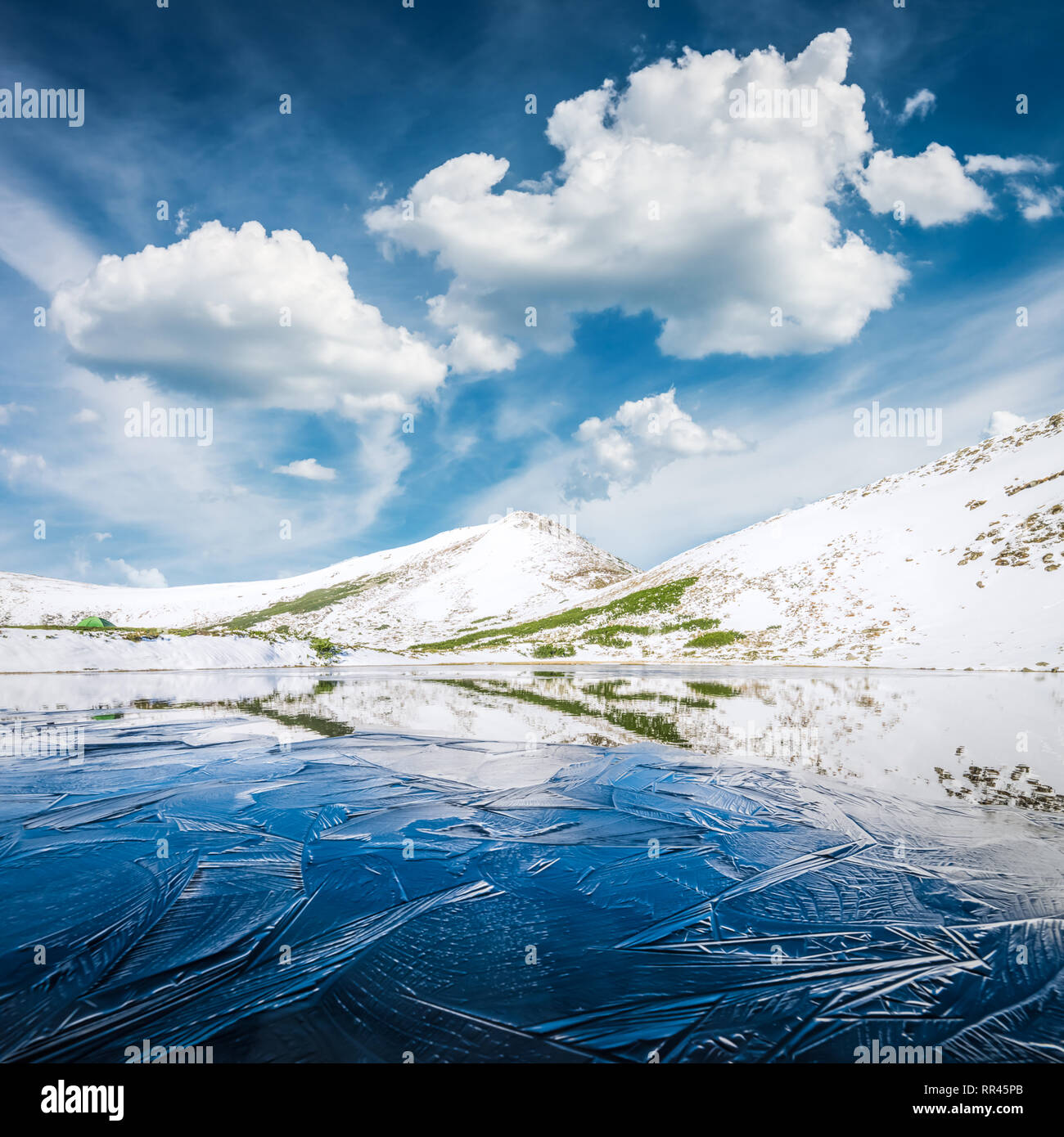 Gefrorenen Bergsee mit Blue Ice und Risse auf der Oberfläche. Malerische Landschaft mit schneebedeckten Hügeln unter einem blauen bewölkten Himmel Stockfoto