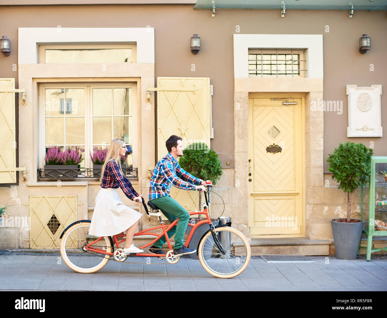 Seitenansicht der aktiven touristische Paar in Freizeitkleidung, bärtigen Mann und die blonde Frau in den Brillen Fahrrad tandem Fahrrad entlang Leer gepflasterten Gehweg auf die hellen sonnigen Sommertag nach Stadt Wohnhäuser. Stockfoto