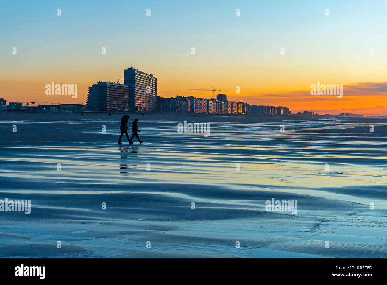 Reflexion von zwei Silhouetten, die auf der Nordsee Strand von Ostende mit urbanen Stadtlandschaft im Hintergrund bei Sonnenuntergang, Westflandern, Belgien. Stockfoto