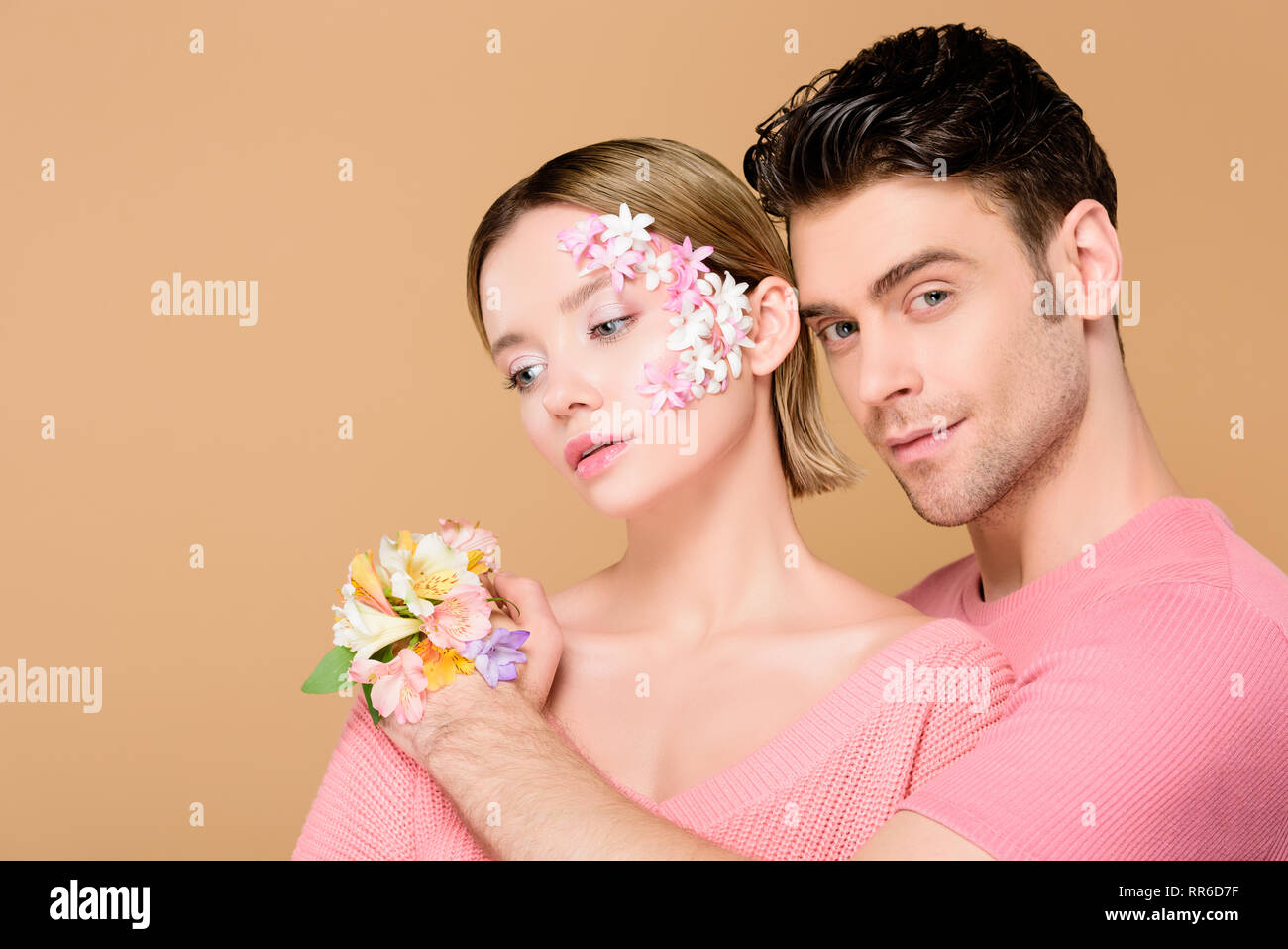 Schöner Freund umarmen Freundin mit Blumen auf dem Gesicht isoliert auf beige Stockfoto Schöner Freund umarmen Freundin mit Blumen auf dem Gesicht isoliert auf beige Stockfoto