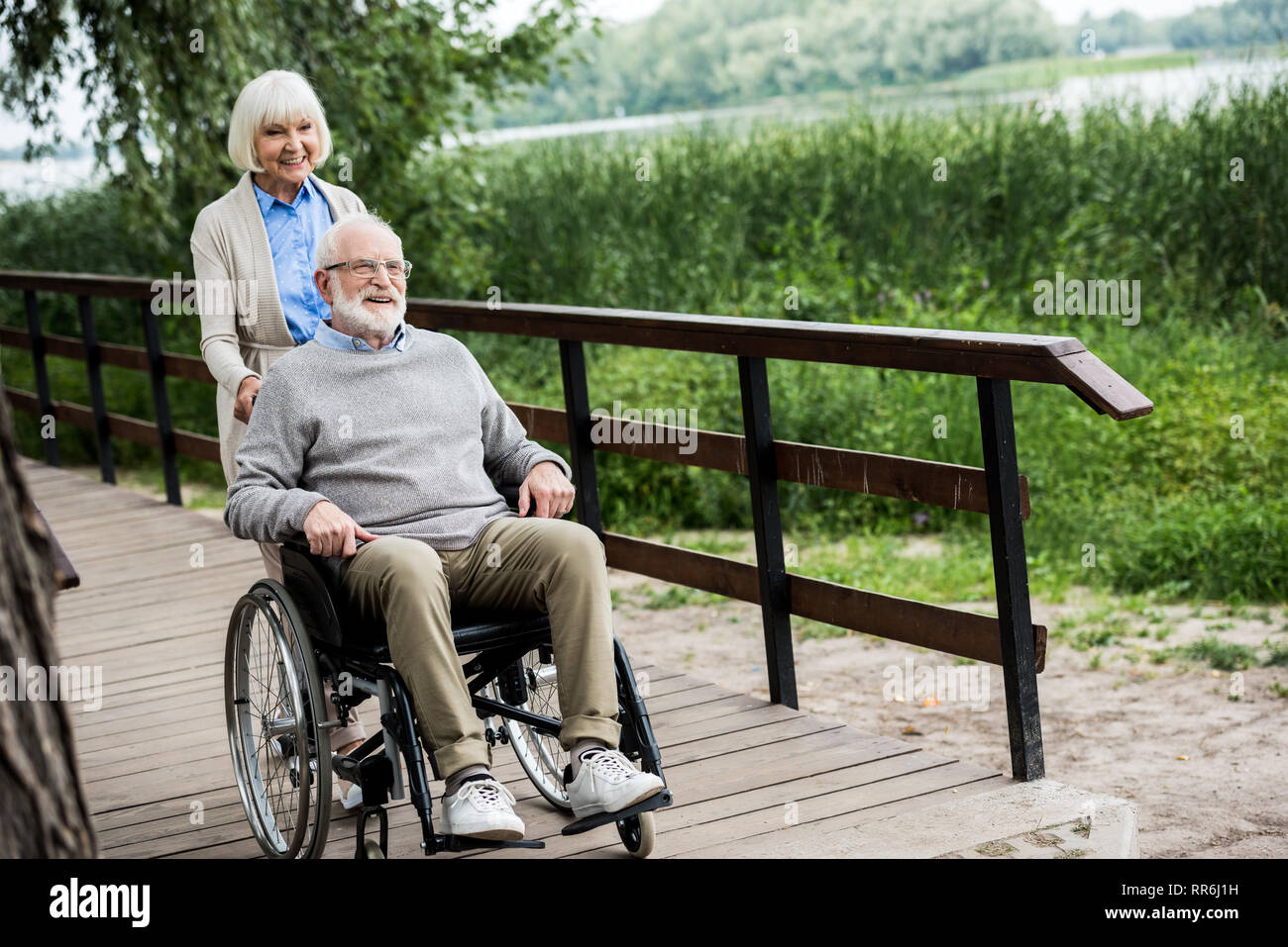 Gerne älterer Mann im Rollstuhl mit lächelnden Frau auf hölzernen Brücke im Park Stockfoto