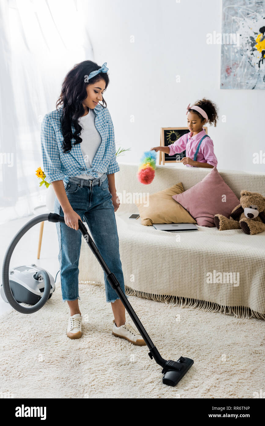 Attraktive afrikanische amerikanische Frau Reinigung Teppich mit Staubsauger und niedlichen Tochter abstauben Sofa Stockfoto