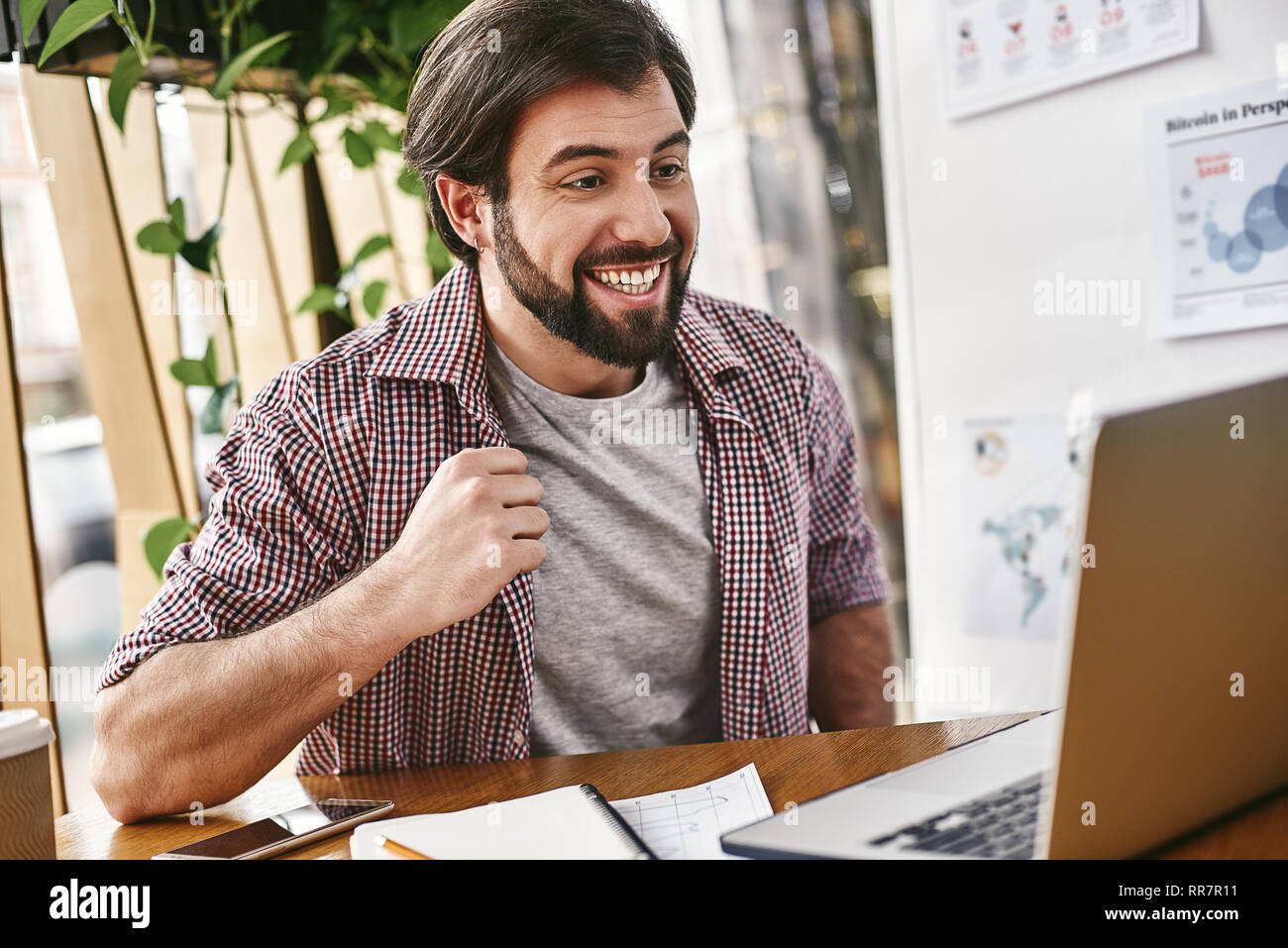Portrait von Stattlichen glücklich bärtige junge Unternehmer in Freizeitkleidung, im Büro und lächelnd, während die Analyse von Statistiken. Er benutzt seinen Laptop, auf dem Bildschirm mit toothy Lächeln Stockfoto
