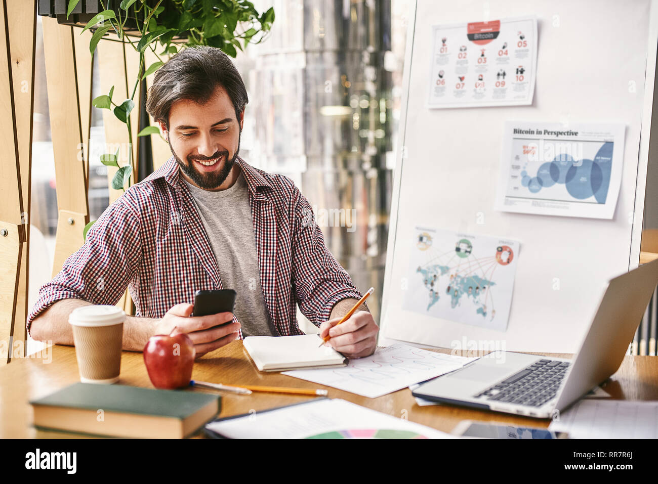 Junge fröhliche Geschäftsmann in Freizeitkleidung ist auf der Suche nach dem Telefon, während im Büro vor dem Laptop, es zu verwenden, Notizen auf Karte. Freelancer analysiert Daten, entwickelt. Business Bildung für Erwachsene. Notebooks, Planer, einen Apfel und eine Tasse Kaffee auf dem Schreibtisch. Stockfoto