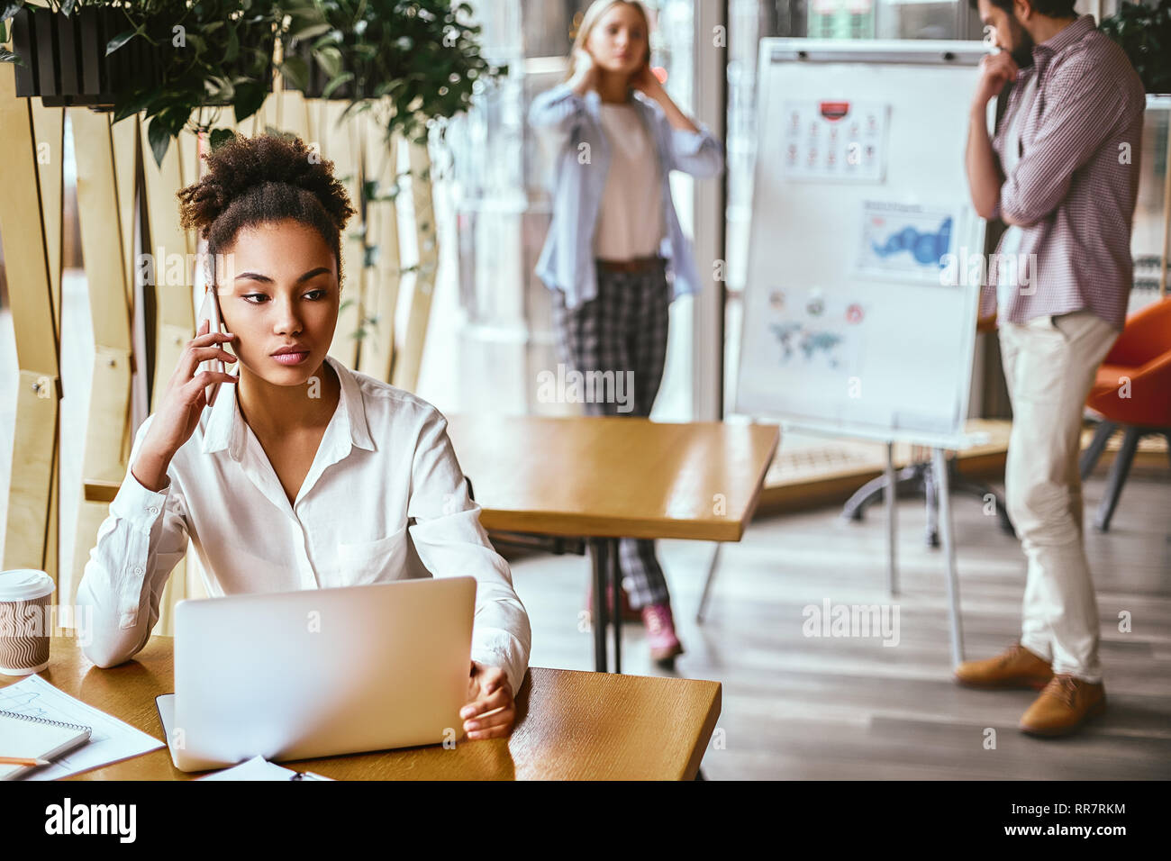 Porträt von Ernst junge Frau an ihrem Schreibtisch sitzen, Gespräch am Handy. Konzentriertes Business woman Gespräch mit Client über Smart Phone im Büro und beiseite. Frau Arbeiten am Schreibtisch in modernen Büro. Ihr Notebook und Kaffee auf den Tisch. Kollegen in den Hintergrund. Stockfoto