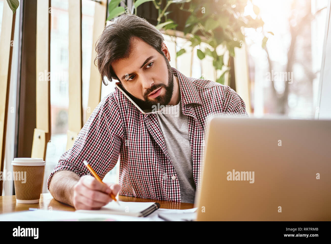 Junge schwere Geschäftsmann in Freizeitkleidung ist, am Telefon zu sprechen, während im Büro vor dem Laptop, es zu verwenden, Notizen auf Karte. Freelancer analysiert Daten, entwickelt. Business Bildung für Erwachsene. Stockfoto