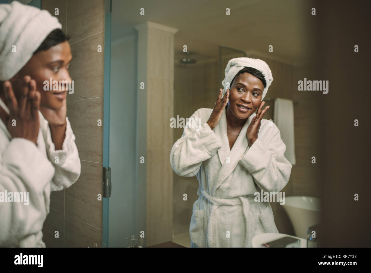 Reife Frau im Bademantel im Badezimmer Spiegel während der Anwendung Feuchtigkeitscreme auf ihrem Gesicht. Afrikanische Frau Anwendung Creme im Gesicht im Bad zu Hause. Stockfoto