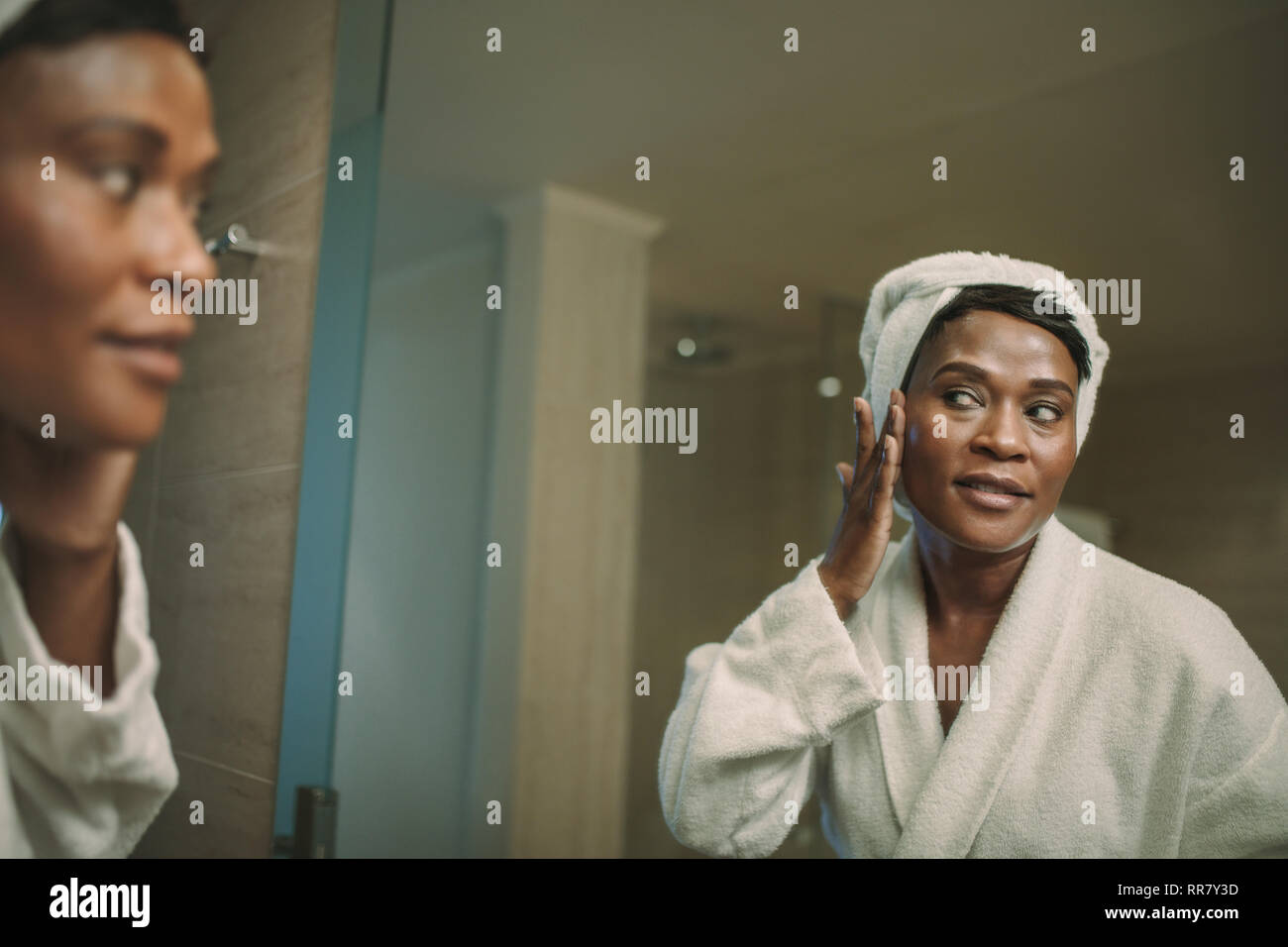 Überlegungen zur Reife der afrikanischen Frau im Badezimmer spiegel Anwendung kosmetische Creme im Gesicht. Reife Frau im Bademantel auf die Sahne auf ihrem Gesicht haut Stockfoto