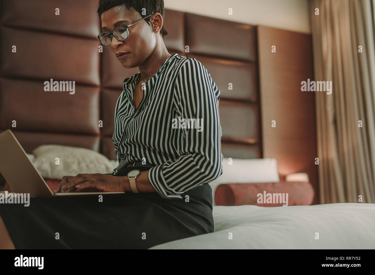 Afrikanische Frau in formelle Kleidung sitzen auf dem Bett im Hotel Zimmer mit Laptop. Geschäftsfrau arbeiten vom Hotelzimmer. Stockfoto