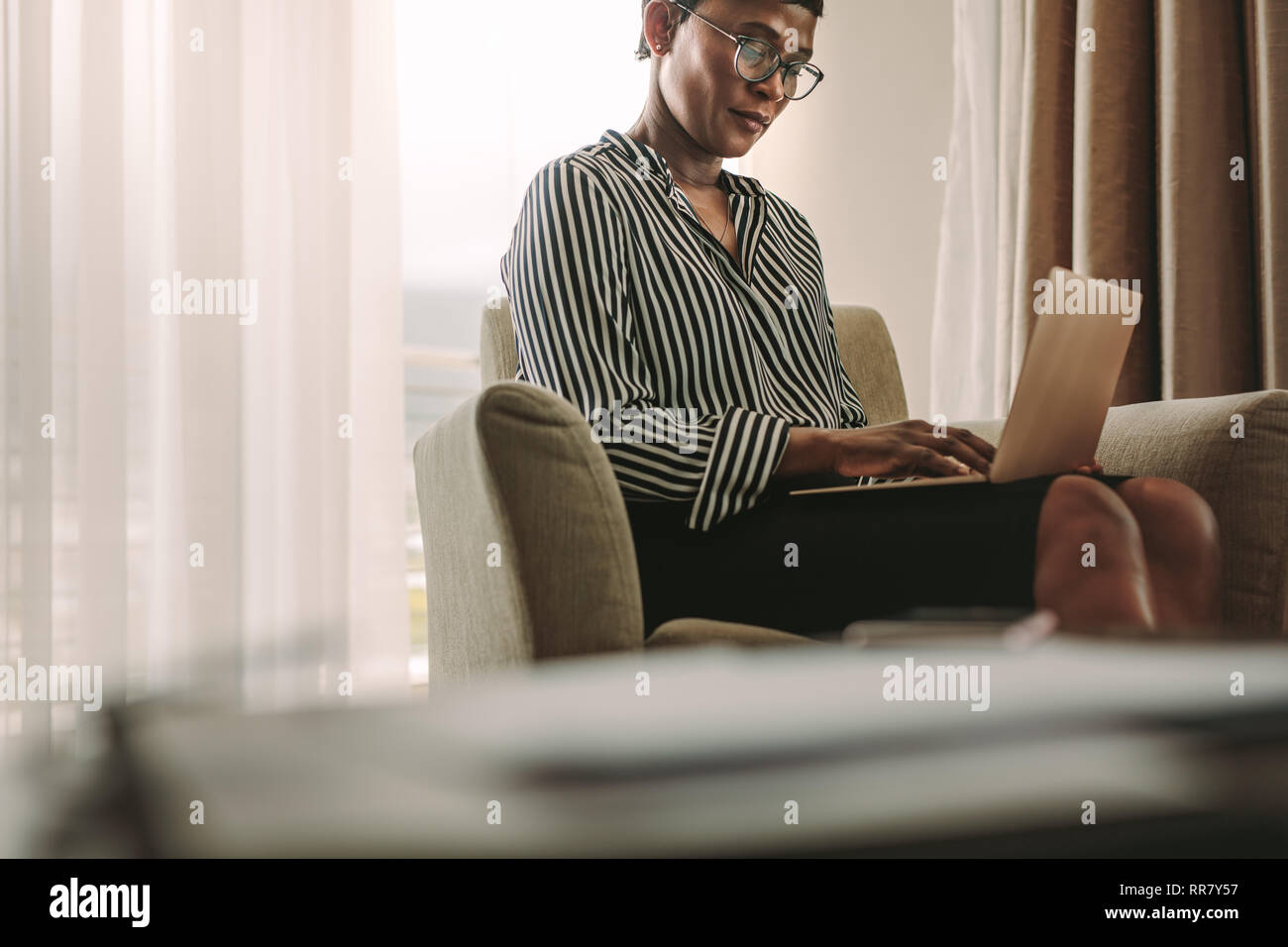 Afrikanische Frau in formelle Kleidung sitzt auf einem Sessel im Hotel Zimmer mit Laptop. Geschäftsfrau auf Geschäftsreise arbeiten vom Hotelzimmer. Stockfoto