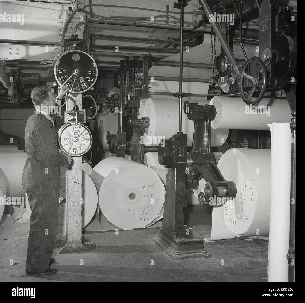 1950er Jahre, männlichen Arbeitnehmer in Latzhosen Kontrolle der Zifferblätter und Bedienelemente in einem Print funktioniert Der brittains Papierfabrik, England, UK, in dem die Maschinen, bei denen die fertigen Papierrollen gelagert werden. Stockfoto