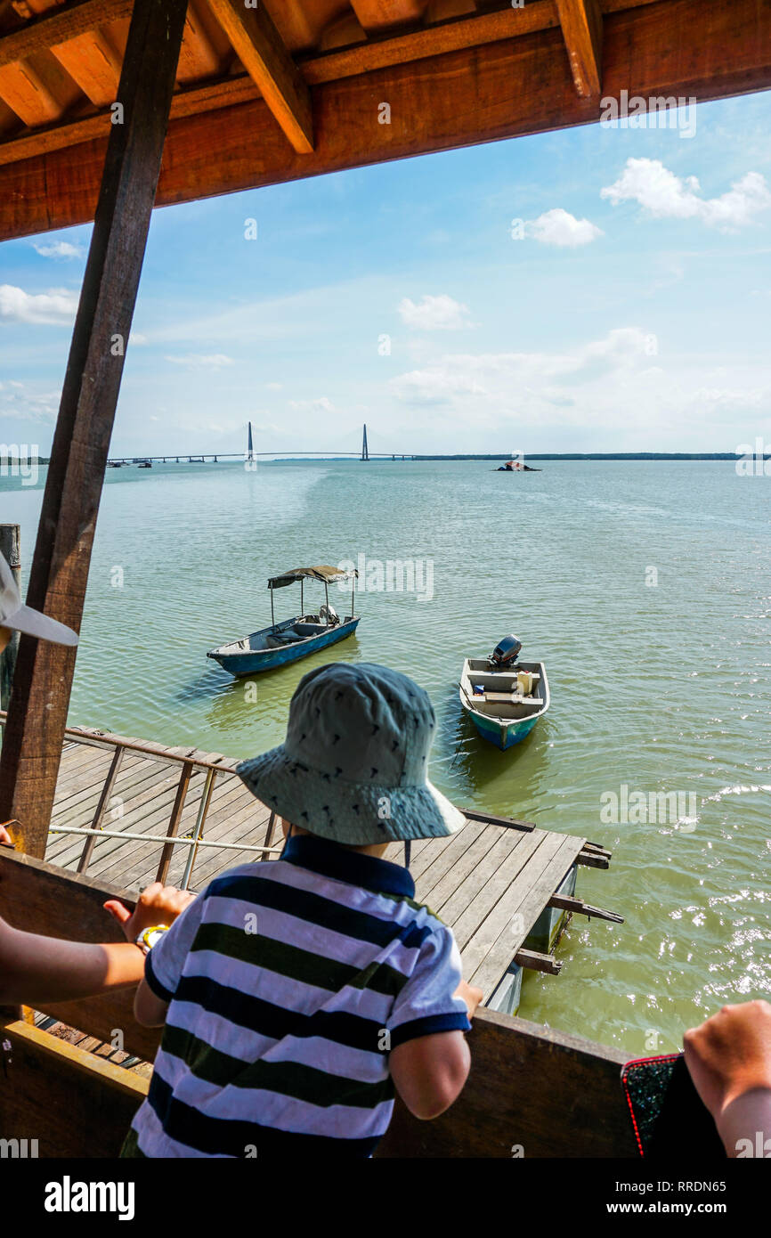 Blick auf unscharfen Vordergrund Kinder beobachten das Boot von den Holzsteg mit Johor River Bridge im Hintergrund angedockt. Blick von einem Holzsteg Stockfoto