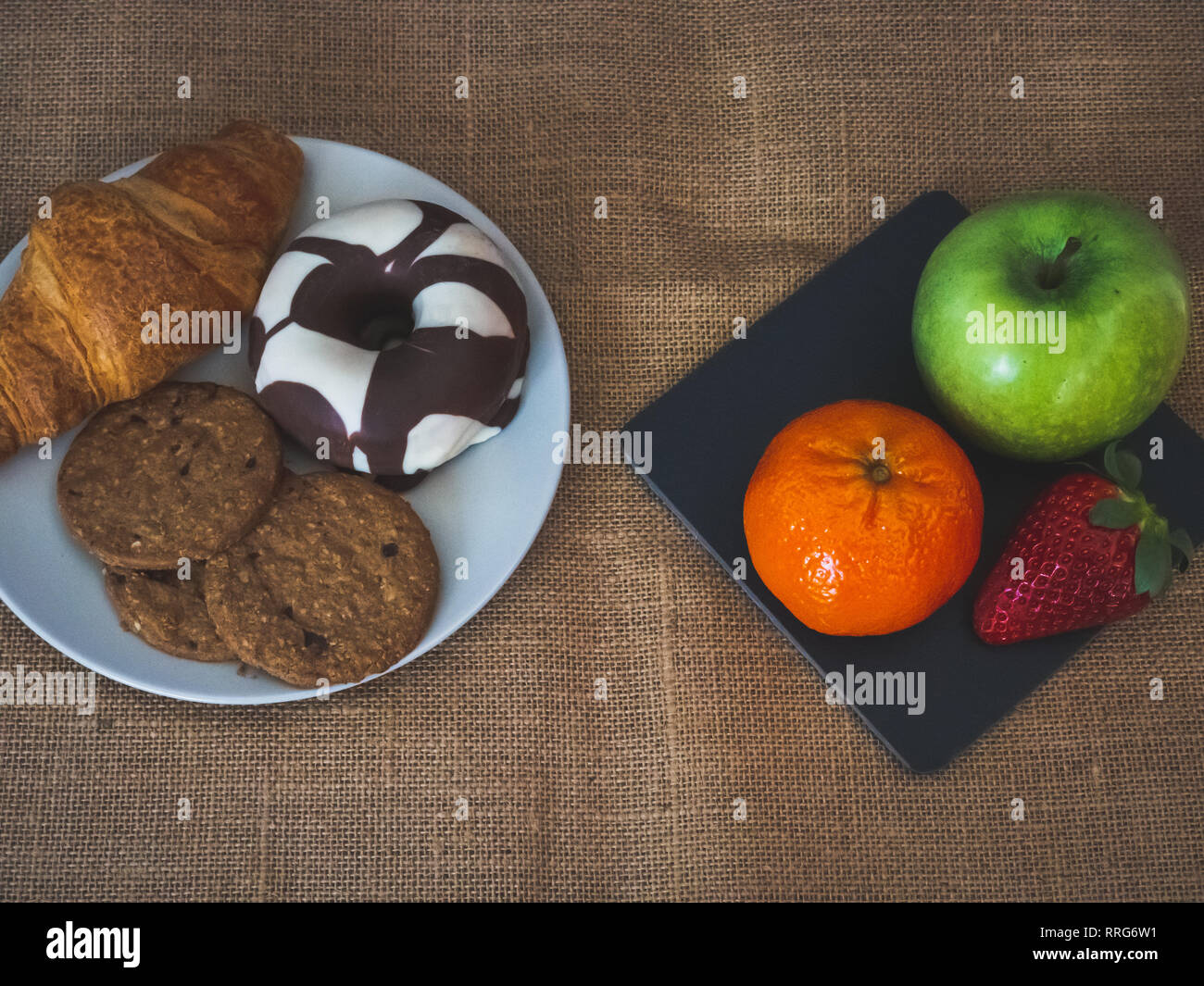 Eine Platte mit schwarzem Schiefer mit einem grünen Apfel, eine Mandarine und eine Erdbeere und eine Platte mit einem Croissant, einem Schokolade Donut und Cookies auf einer Leinwand Stockfoto