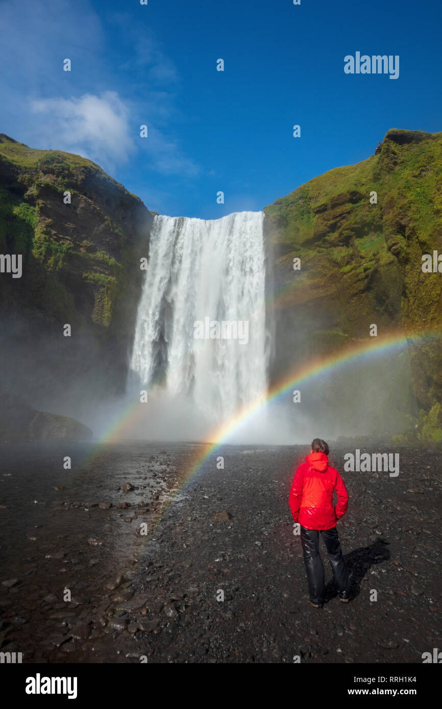 Rainbow und Person unter 60 m hohen Wasserfall Skogafoss. Skogar, Sudhurland, South Island. Stockfoto