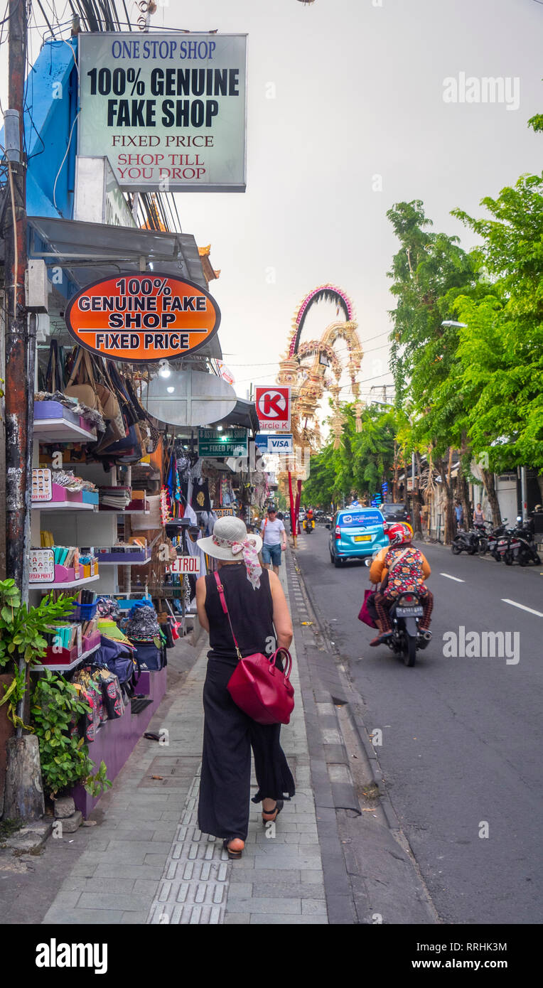 Kaukasische weibliche Touristen mit roten Tasche vorbei eine humorvolle Zeichen von 100% Echtes Fake Shop auf Jl Raya Legian, Kuta Bali Indonesien. Stockfoto
