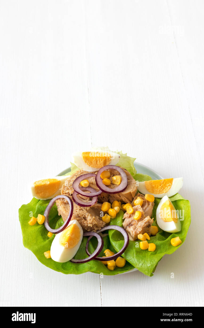 Thunfisch in Dosen mit Eiern, suggar Mais, rote Zwiebel Ringe auf grünen Salatblatt, Scheibe Zitrone in pocelain Schüssel. Thunfisch Salat. Weiße Holztisch backgr Stockfotografie