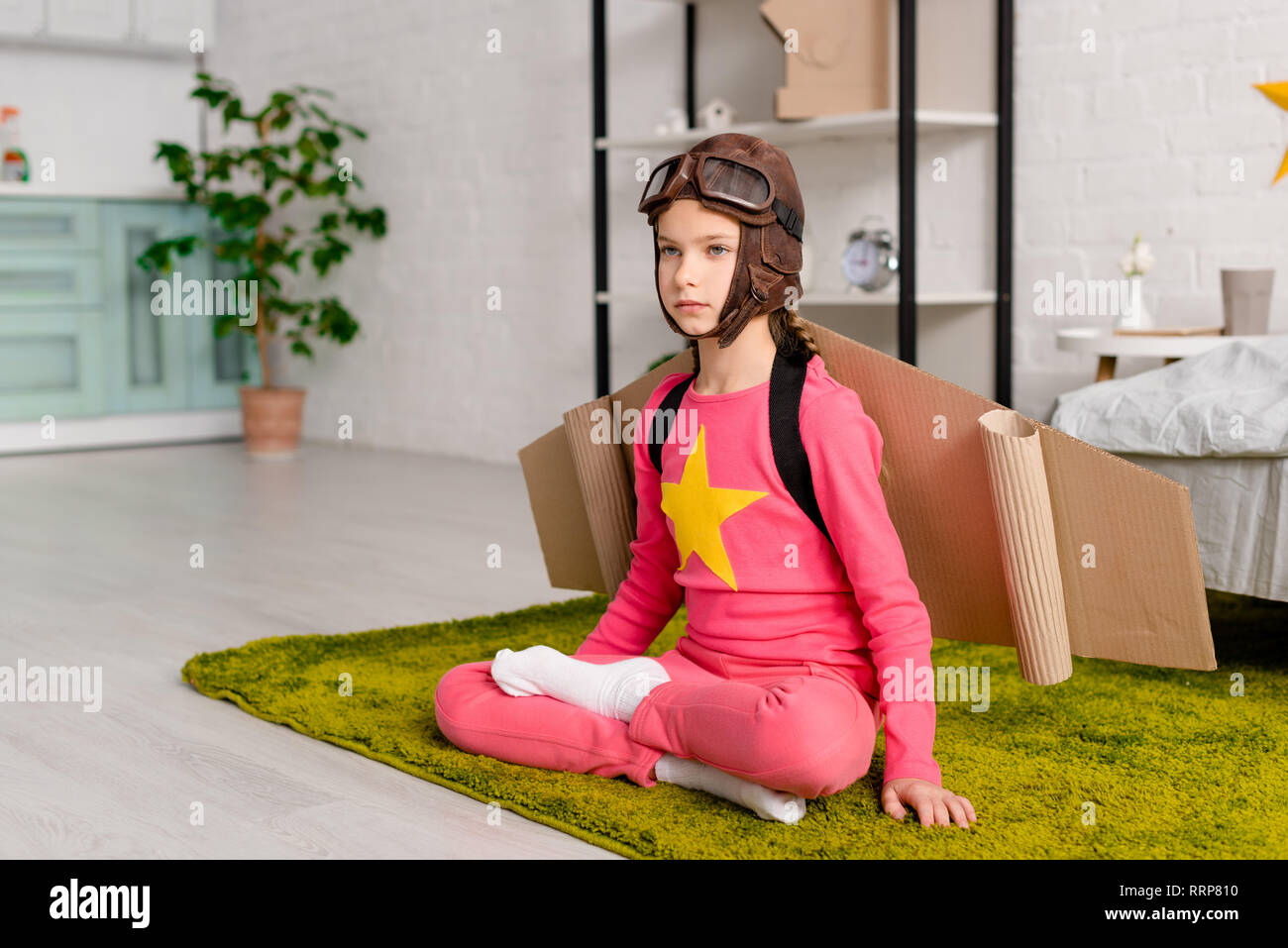 Schwere Kind mit Karton Flügeln sitzt im Lotussitz auf dem Teppich Stockfoto