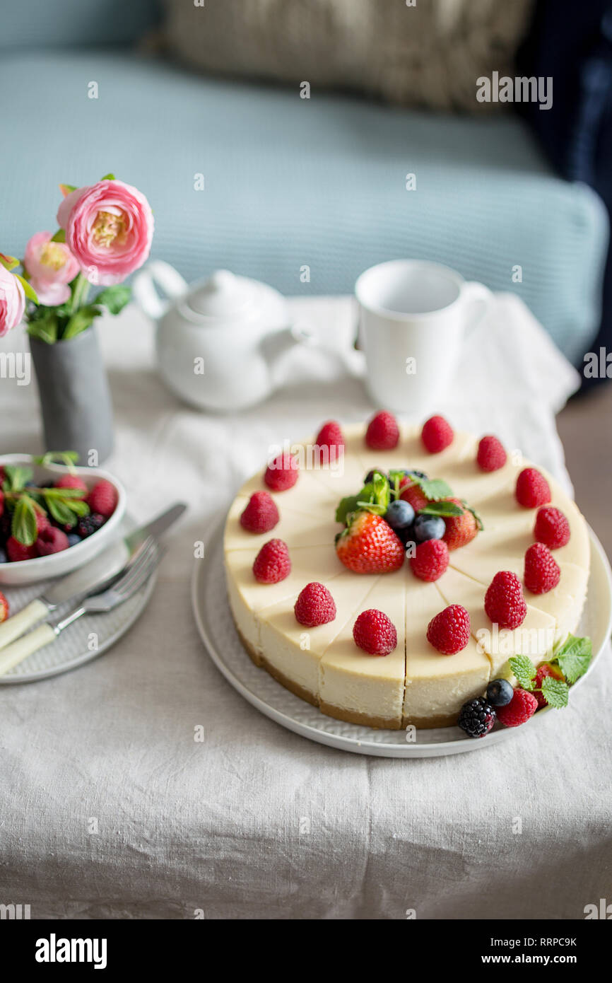 Käsekuchen, hausgemachten Kuchen mit Erdbeeren Beeren in eine graue Platte, mit Schale, Teekanne, Gabel, Messer, frische Beeren, Vase mit Blumen, Seitenansicht. Süßigkeiten Stockfoto