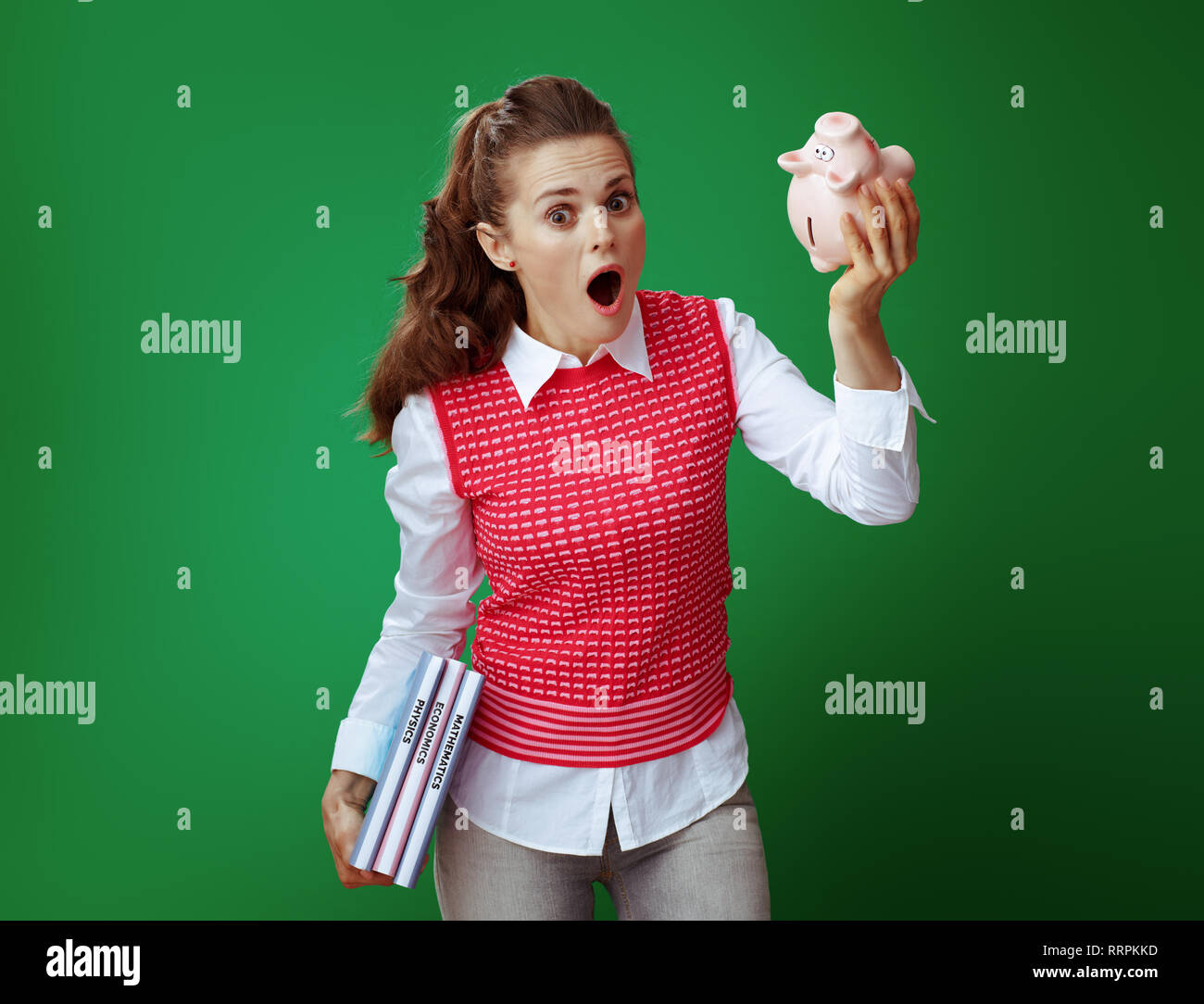 Moderne Student in Grau jeans und Pink ärmelloses Shirt mit Lehrbüchern schütteln rosa Sparschwein vor grünem Hintergrund schockiert. Finanzielle Herausforderungen a Stockfoto