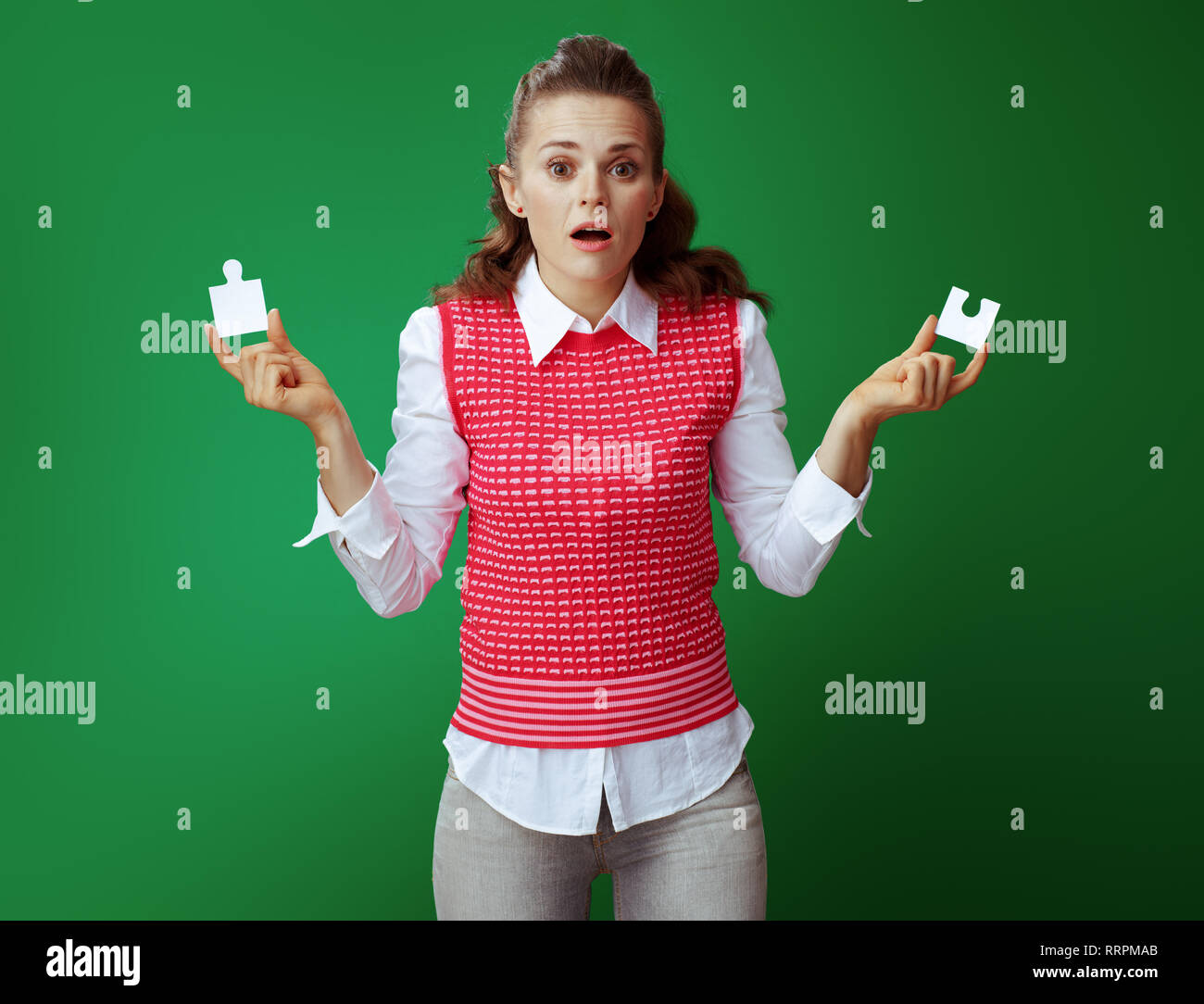 Verwirrt moderne Lerner Frau in Grau jeans und Pink ärmelloses Shirt mit 2 Stück des Puzzles isoliert auf Grün. Mangelndes analytisches Denken, re Stockfoto