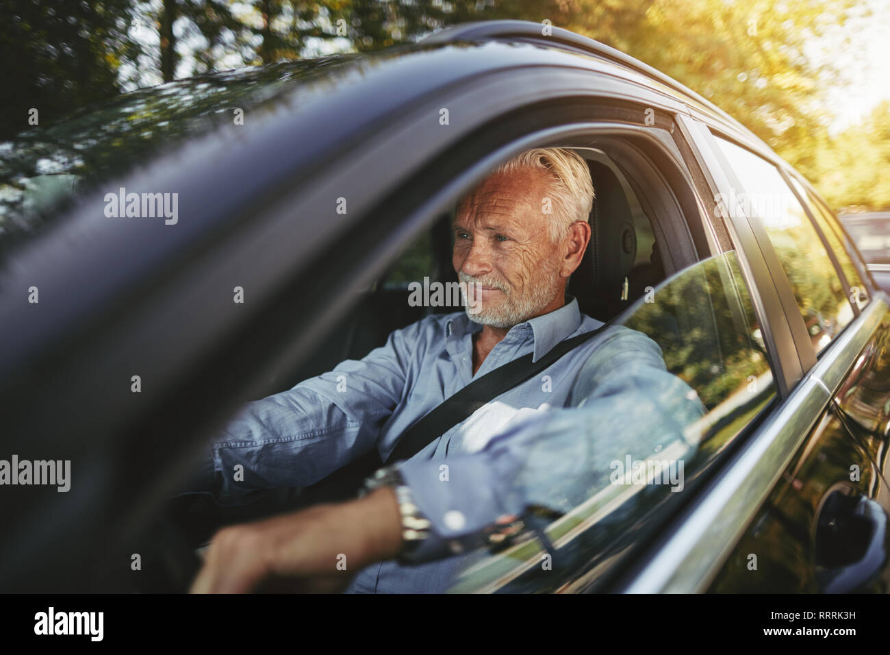 Älterer Mann lächelnd, während allein in seinem Auto fahren entlang einer von Bäumen gesäumten Straße im Land Stockfoto