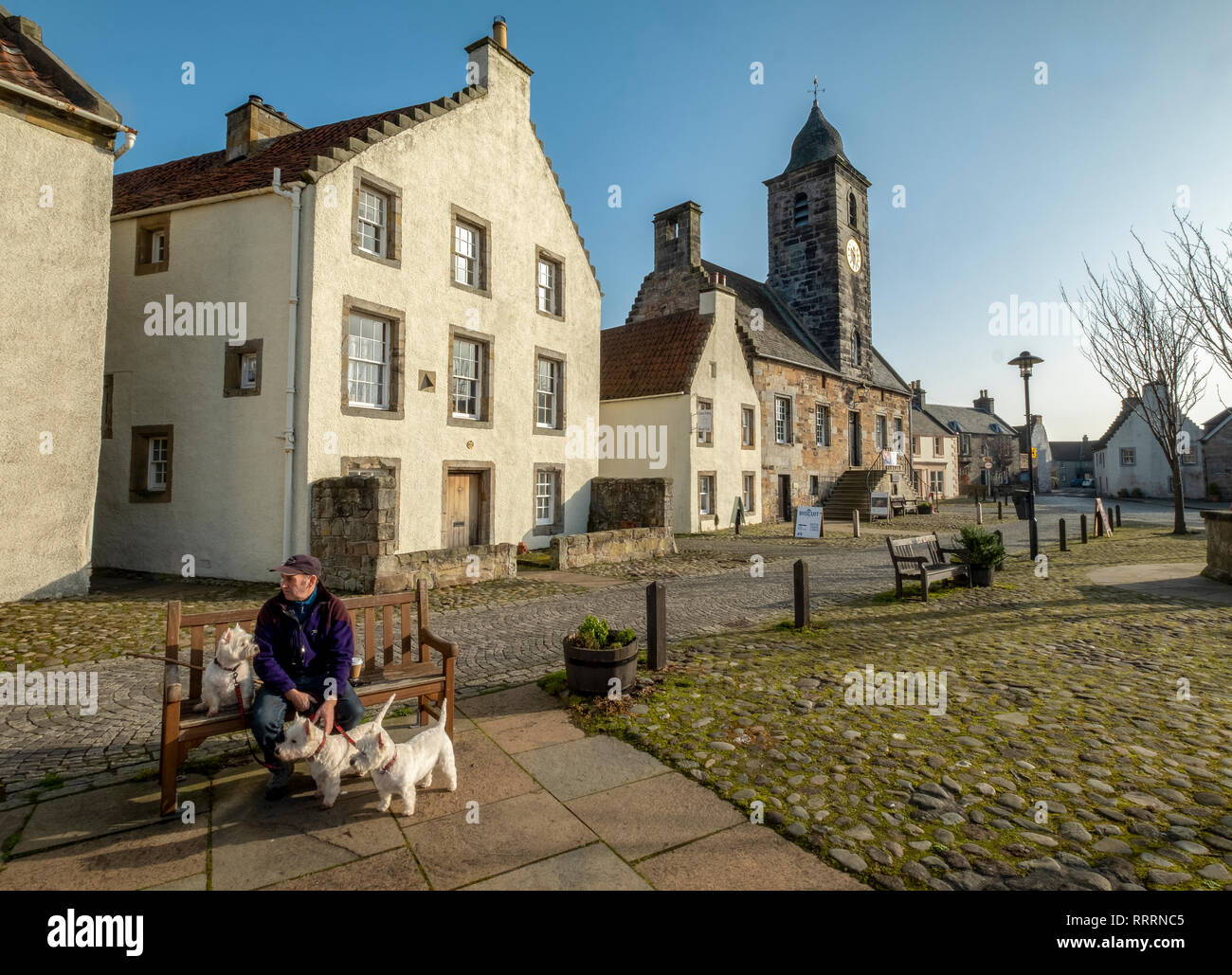 Ein Mann mit drei West Highland Terrier sitzen auf einer Bank in dem historischen Dorf folgende Sehenswürdigkeiten: Culross, Fife, Schottland. Stockfoto