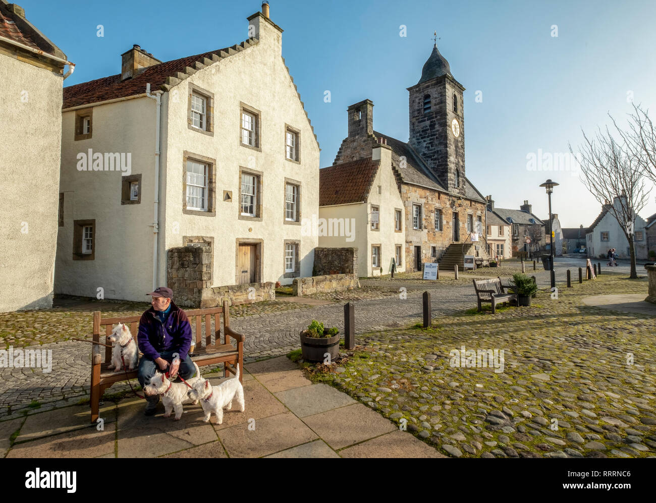 Ein Mann mit drei West Highland Terrier sitzen auf einer Bank in dem historischen Dorf folgende Sehenswürdigkeiten: Culross, Fife, Schottland. Stockfoto