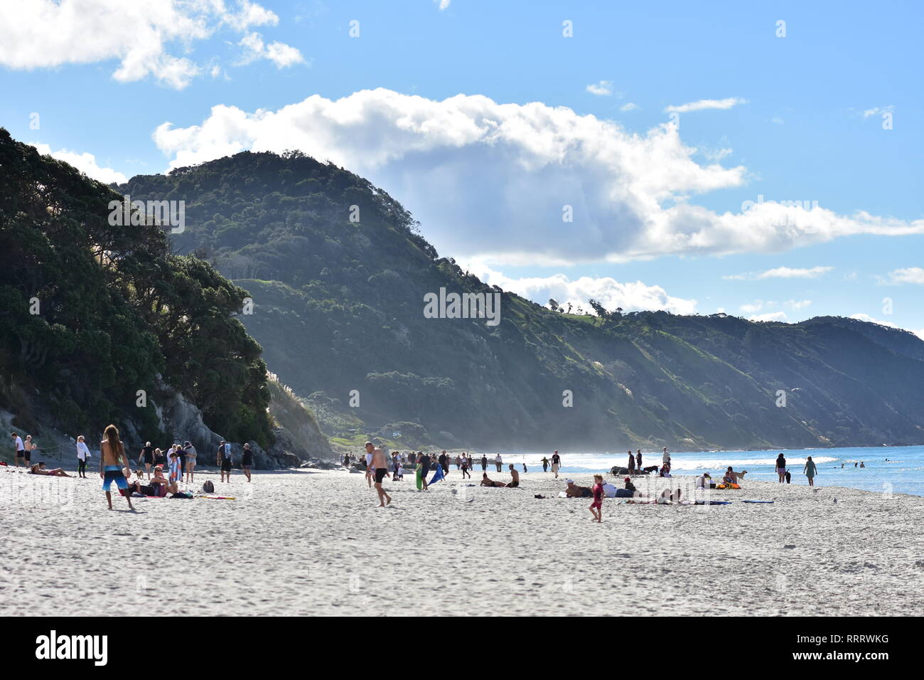 Massen von Menschen genießen, sonnigen Tag auf einem flachen Sandstrand Pacific Ocean Beach in Mangawhai Heads mit steilen Hügeln im Hintergrund. Stockfoto