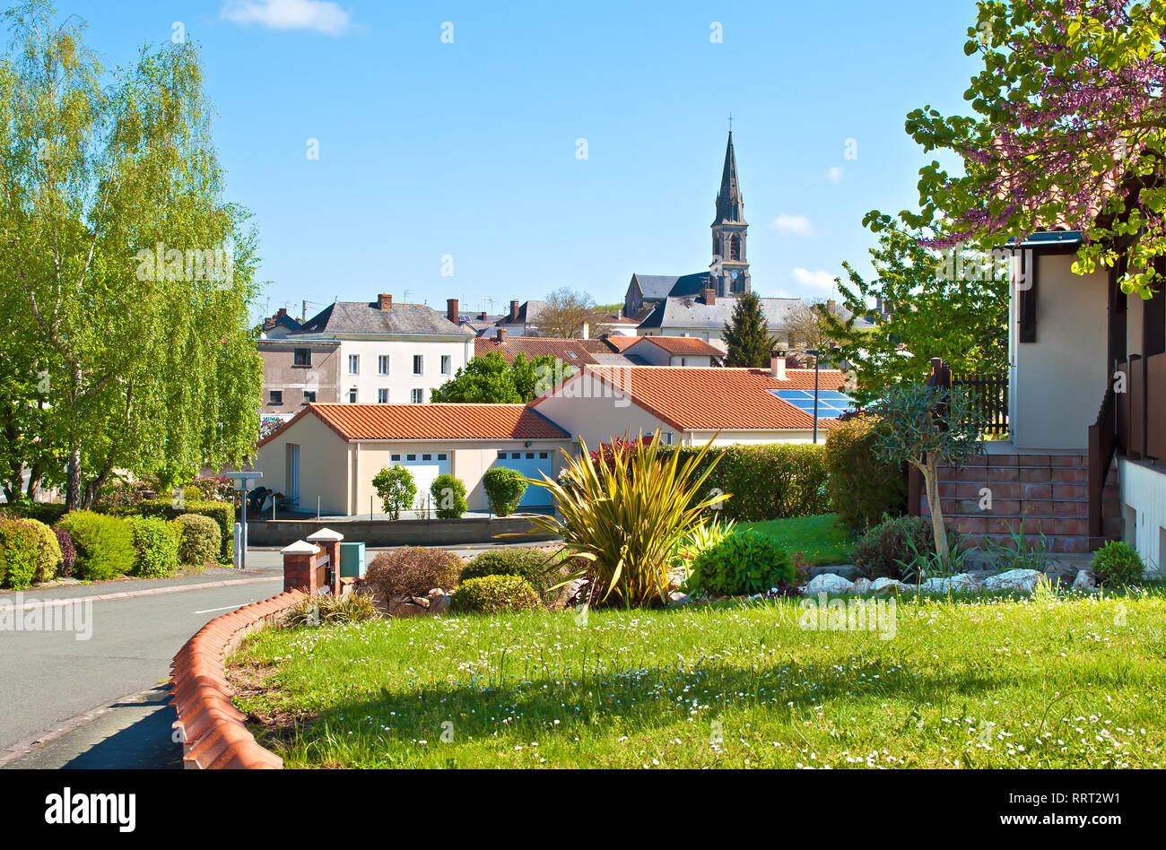 Blick auf eine kleine französische Stadt Neuvy-en-Mauges, Frankreich. Viele Häuser unter grünen Bäumen und die roten Dächer mit zellulären Panels, Turm einer Kirche. Warme spri Stockfoto