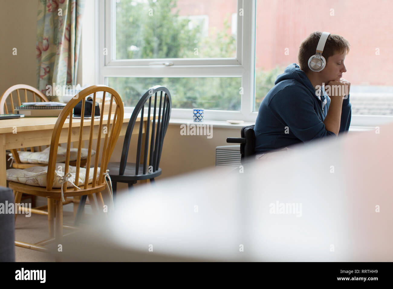 Ruhige junge Frau im Rollstuhl Musikhören mit Kopfhörern mit Fenster Stockfoto