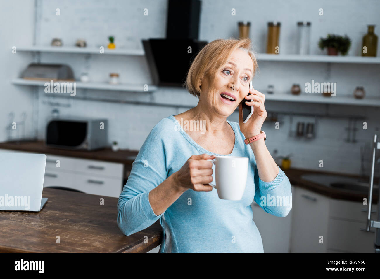 Überrascht ältere Frau mit Tasse Kaffee sprechen auf dem Smartphone zu Hause Stockfoto